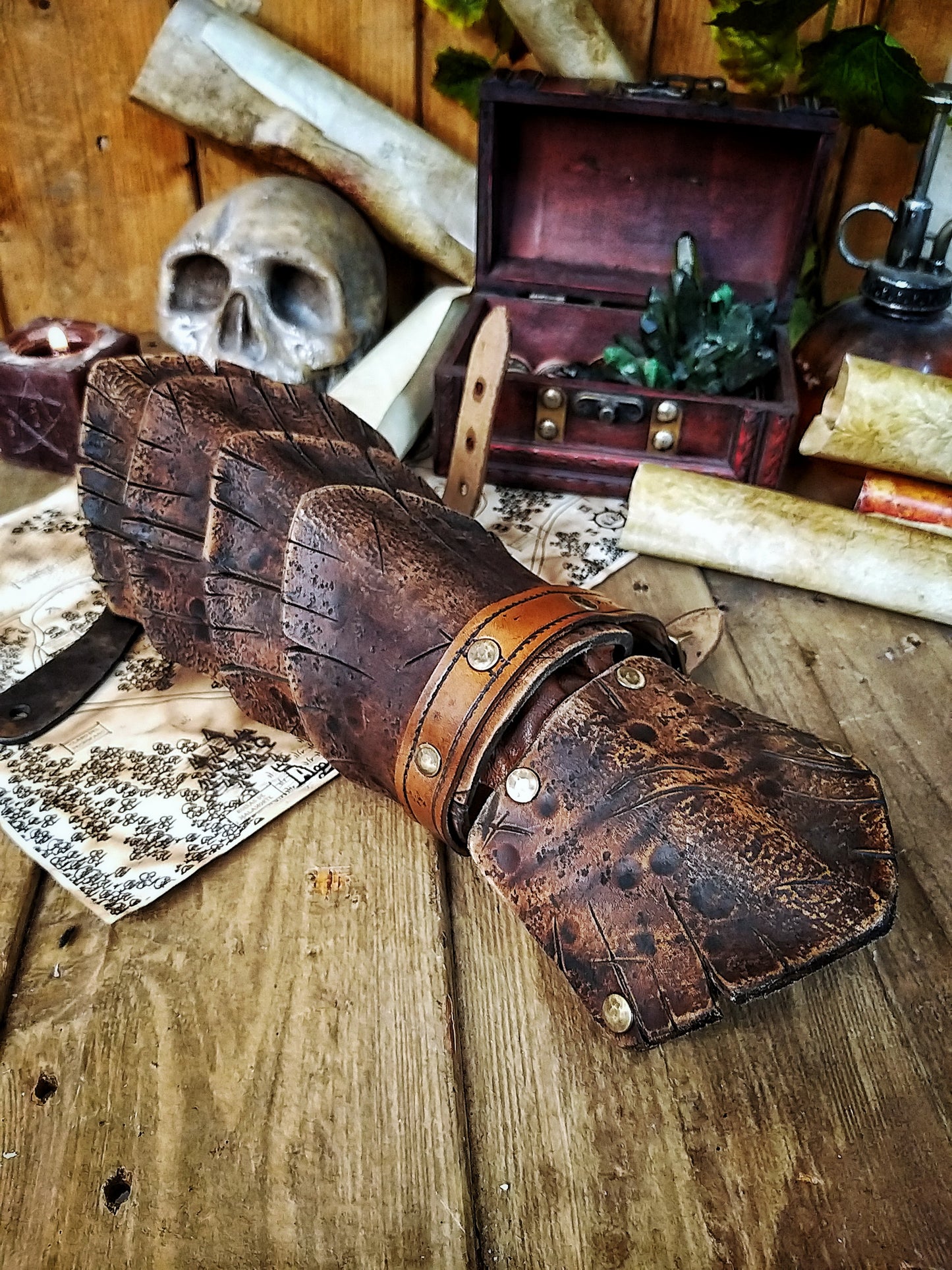 Dragon Scale Leather Bracer: LARP Cosplay Armor for Fantasy Warriors, Orcs, Archers - Handmade Medieval Costume A brown leather dragon scale bracer displayed on a wooden surface, with a skull and vintage-style props in the background, suggesting a medieval or fantasy theme.