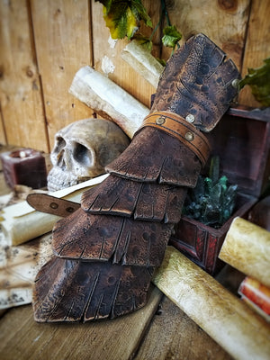 A brown handmade Dragon Scales Leather Bracer is worn on a man’s forearm, fully fastened and resting over a vintage map surrounded by fantasy props like a skull and candle. The layered leather plates resemble dragon hide, with an aged finish and rugged style. Perfect for LARP warriors, monster hunters or medieval cosplay. The leather is vegetable-tanned and hand-painted using Outcast Props’ signature weathering technique.