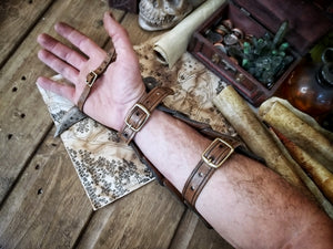 Close-up of a male arm wearing an open Dragon Scales Leather Bracer, showing the inner leather straps and antique brass buckles used to fasten it. The bracer lies on a rustic wooden table with medieval props like a map, coins, scrolls and a skull. Ideal for LARP warrior costume, fantasy fair outfits or orc archer cosplay. This handcrafted bracer is made of premium vegetable-tanned leather using traditional Spanish techniques.