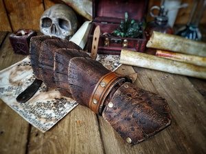 A brown handmade Dragon Scales Leather Bracer is worn on a man’s forearm, fully fastened and resting over a vintage map surrounded by fantasy props like a skull and candle. The layered leather plates resemble dragon hide, with an aged finish and rugged style. Perfect for LARP warriors, monster hunters or medieval cosplay. The leather is vegetable-tanned and hand-painted using Outcast Props’ signature weathering technique.