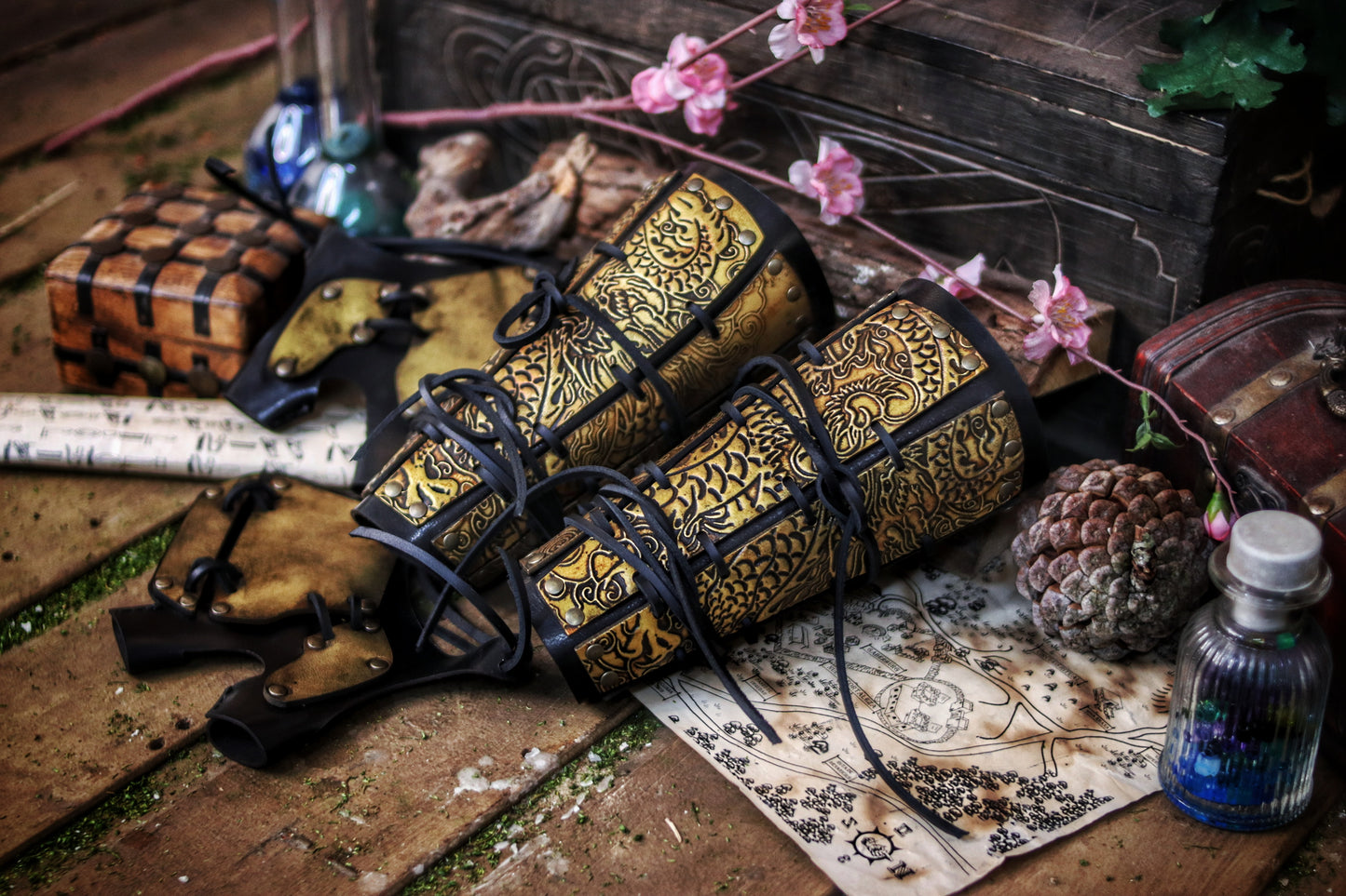 Golden engraved samurai armor bracers resting on a table with fantasy props like potions and maps. Designed for samurai larp costume and cosplay warriors. Black lacing and ornate dragon motifs give a powerful Eastern feel. Durable and handcrafted with vegetable-tanned leather and water-based dyes. Aged to resemble antique Japanese armor pieces. Great for L5A RPG or fantasy samurai characters in immersive games.