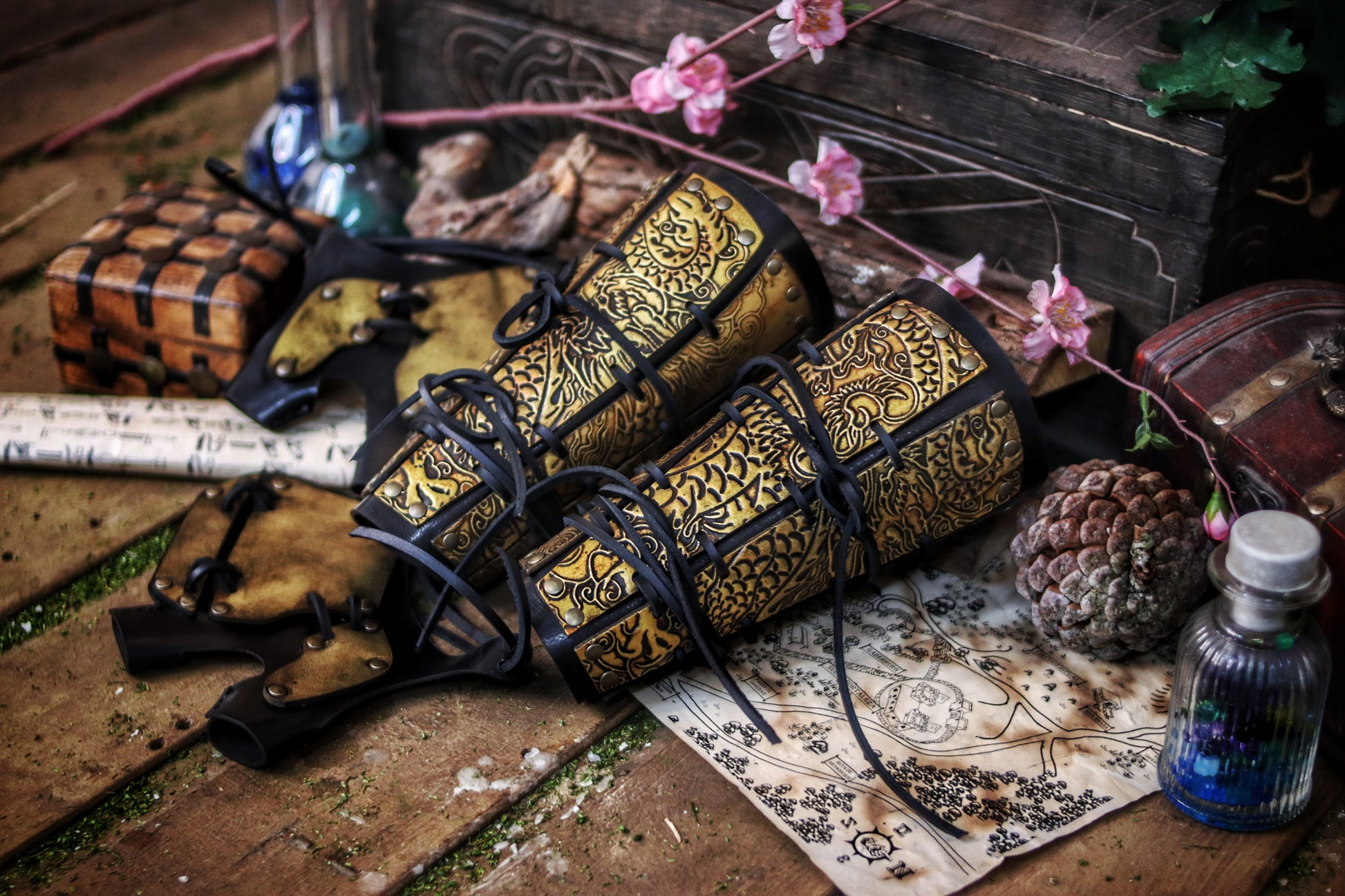 Golden engraved samurai armor bracers resting on a table with fantasy props like potions and maps. Designed for samurai larp costume and cosplay warriors. Black lacing and ornate dragon motifs give a powerful Eastern feel. Durable and handcrafted with vegetable-tanned leather and water-based dyes. Aged to resemble antique Japanese armor pieces. Great for L5A RPG or fantasy samurai characters in immersive games.