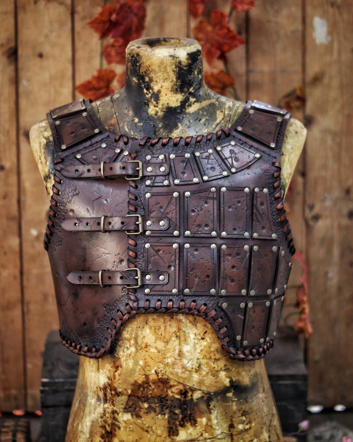A brown leather studded armor piece displayed on a mannequin, featuring chest plates with studs and an adjustable buckle system, with a backdrop of autumn leaves. A handmade leather studded armor displayed on a wooden stand with a fur cape, featuring brown leather plates with studs, and a corded edge finish. Barbarian studded leather chest armor with rabbit fur cape attachment shown on mannequin. Perfect for fantasy LARP, Viking reenactment or cosplay, this full-grain leather armor is aged by hand and paire