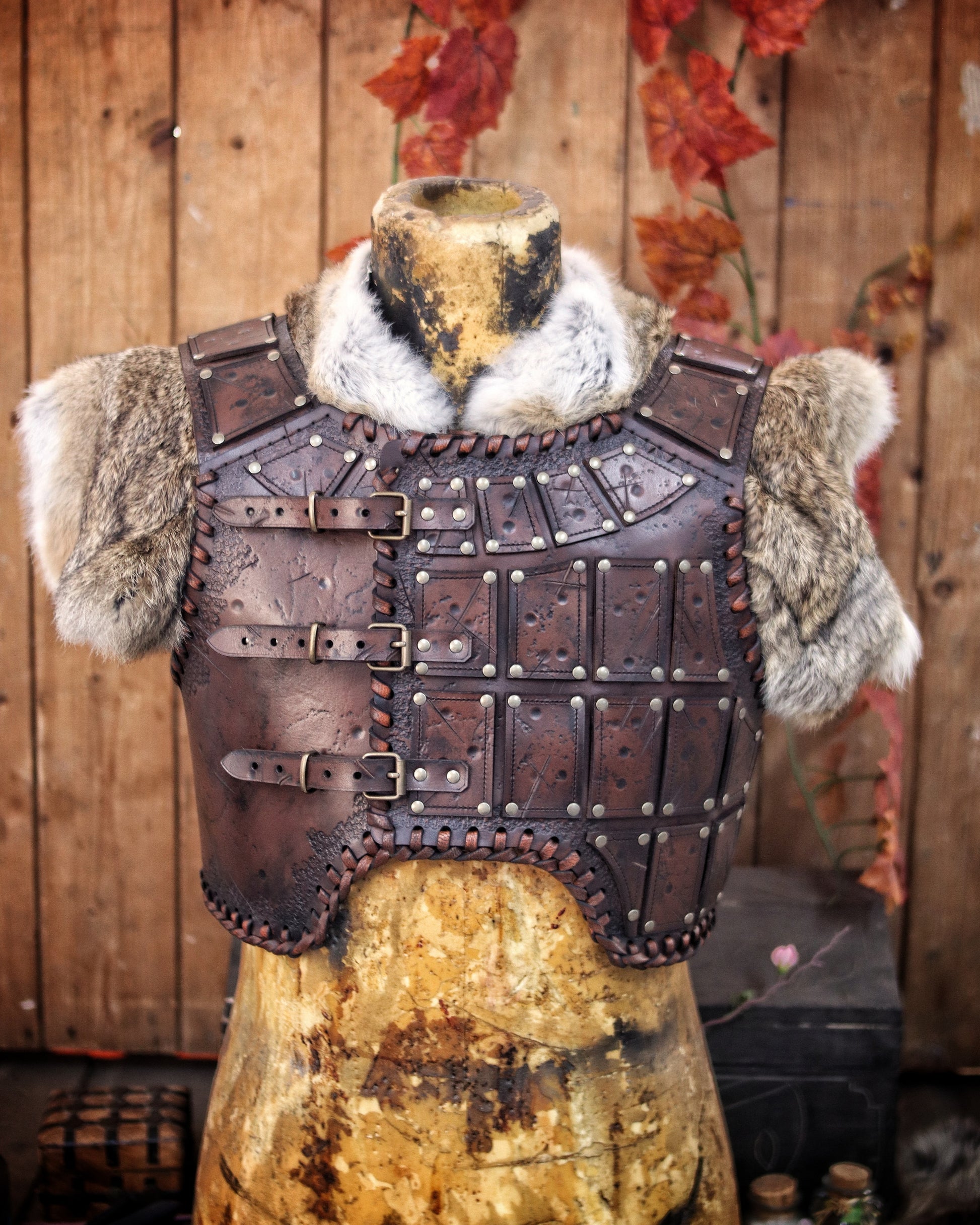 A handmade leather studded armor displayed on a stand, featuring chest plates with studs and an adjustable buckle system, with a fur cape on top. The armor has a brown distressed finish and is positioned against a backdrop with autumn leaves. A handmade leather studded armor displayed on a wooden stand with a fur cape, featuring brown leather plates with studs, and a corded edge finish. Barbarian studded leather chest armor with rabbit fur cape attachment shown on mannequin. Perfect for fantasy LARP, Viking