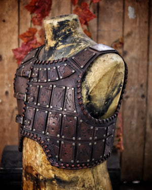 A handmade leather studded armor displayed on a wooden stand with a fur cape, featuring brown leather plates with studs, and a corded edge finish. Barbarian studded leather chest armor with rabbit fur cape attachment shown on mannequin. Perfect for fantasy LARP, Viking reenactment or cosplay, this full-grain leather armor is aged by hand and paired here with fur for an epic look. Adjustable buckle straps and visible stitching add to the rugged charm of this high-impact warrior costume piece.
