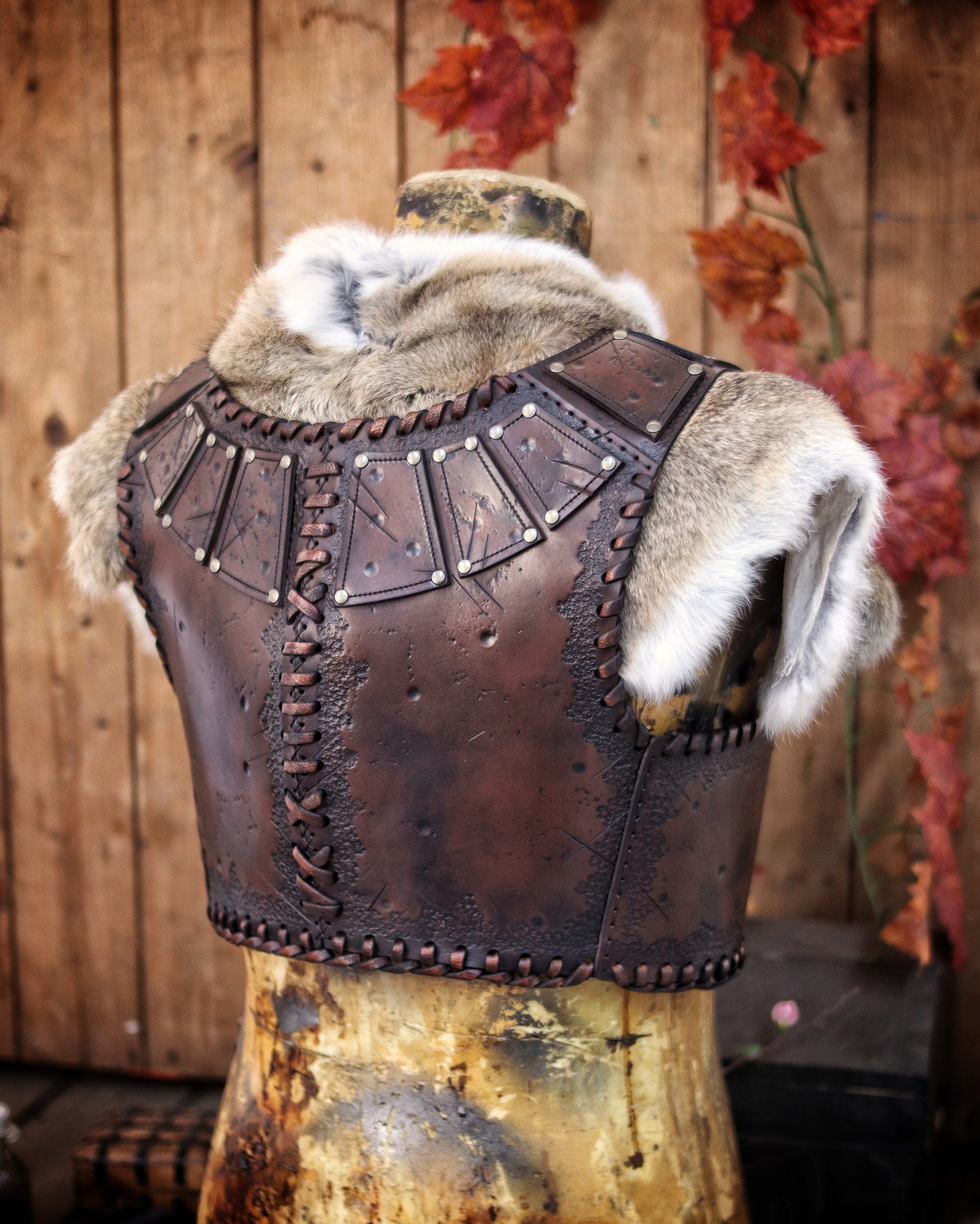 A handmade leather studded armor displayed on a wooden stand with a fur cape, featuring brown leather plates with studs, and a corded edge finish. Barbarian studded leather chest armor with rabbit fur cape attachment shown on mannequin. Perfect for fantasy LARP, Viking reenactment or cosplay, this full-grain leather armor is aged by hand and paired here with fur for an epic look. Adjustable buckle straps and visible stitching add to the rugged charm of this high-impact warrior costume piece.