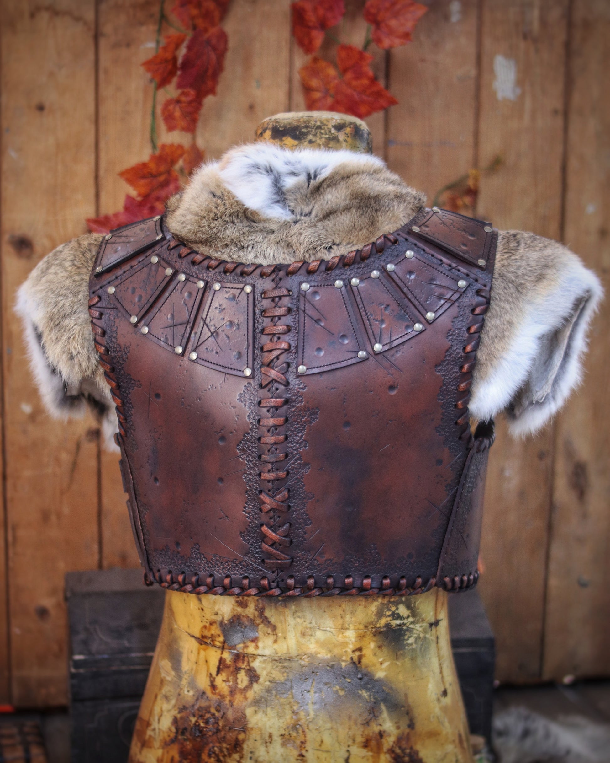 A handmade leather studded armor displayed on a wooden stand with a fur cape, featuring brown leather plates with studs, and a corded edge finish. Barbarian studded leather chest armor with rabbit fur cape attachment shown on mannequin. Perfect for fantasy LARP, Viking reenactment or cosplay, this full-grain leather armor is aged by hand and paired here with fur for an epic look. Adjustable buckle straps and visible stitching add to the rugged charm of this high-impact warrior costume piece.