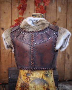 A handmade leather studded armor displayed on a wooden stand with a fur cape, featuring brown leather plates with studs, and a corded edge finish. Barbarian studded leather chest armor with rabbit fur cape attachment shown on mannequin. Perfect for fantasy LARP, Viking reenactment or cosplay, this full-grain leather armor is aged by hand and paired here with fur for an epic look. Adjustable buckle straps and visible stitching add to the rugged charm of this high-impact warrior costume piece.