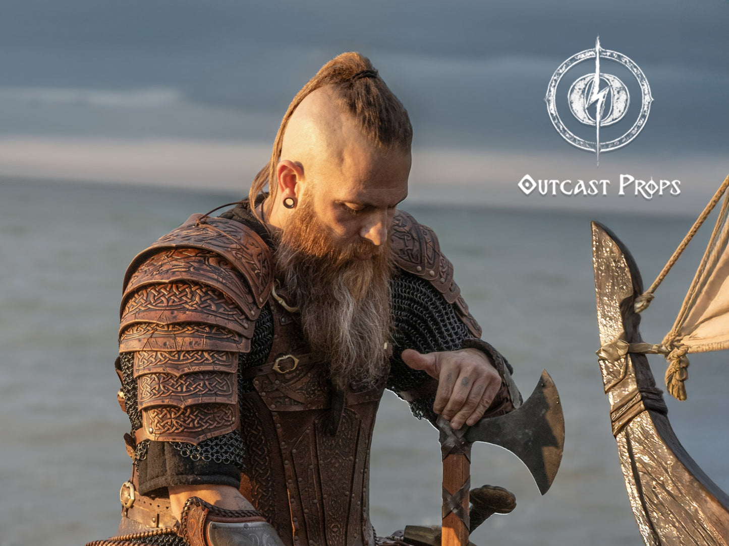 Viking leather shoulder armor with Celtic knot engravings worn by a Viking warrior at the shore, resting his hands on a Viking axe, gazing solemnly. The handcrafted armor features articulated pauldrons for full mobility and is finished with cinematic ageing, perfect for Viking cosplay, LARP shoulder armor, Nordic warrior costume, leather pauldrons, and historical reenactment. The Outcast Props logo is visible on the image.