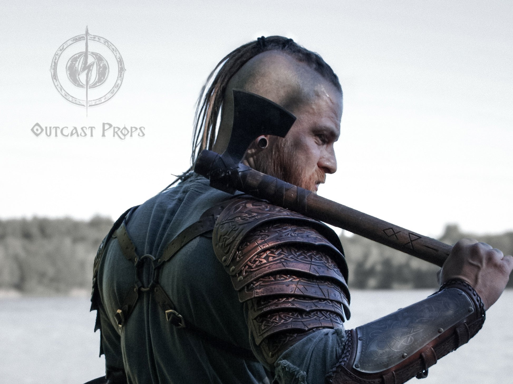 Viking leather shoulder armor with engraved Celtic knot pauldrons seen from behind on a Viking warrior holding a large axe over his shoulder. The handcrafted armor shows detailed leatherwork, flexible construction, and battle-worn finish, ideal for LARP leather armor, Viking cosplay, Nordic warrior shoulder armor, fantasy pauldrons, and medieval reenactment. Outcast Props logo and forested river background complete the scene.