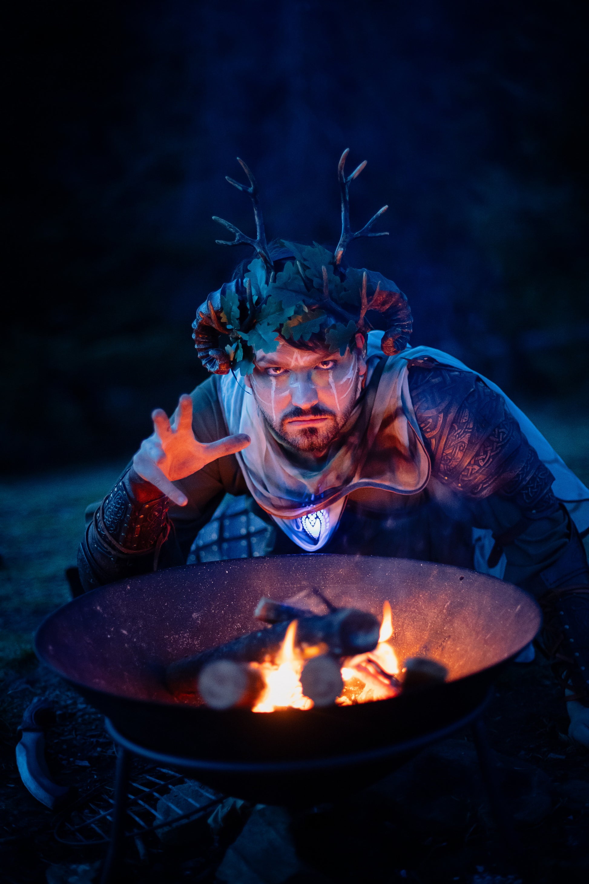 Fantasy druid costume with antler crown, oak leaves, and handcrafted Viking leather shoulder armor Sedrik, richly engraved with Celtic knots for LARP fantasy druid cosplay, magical shaman outfit or Nordic forest mage attire. The druid kneels by a campfire under a blue night sky, performing a ritual. Perfect for LARP, Celtic fantasy costume, druid cosplay, Viking fantasy warrior outfit, shaman costume, and medieval forest events or photoshoots. High-quality leather armor detail.