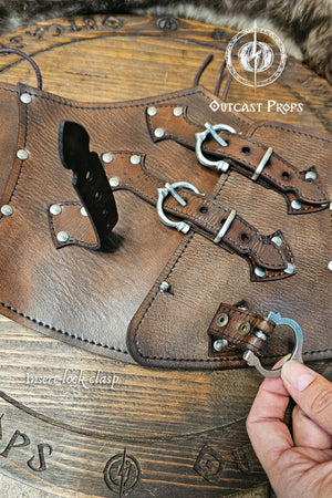 Detailed image of the aged brown leather corset with nickel buckles showing the insert-lock clasp system on a wooden Outcast Props display. The handcrafted underbust corset features triple front straps for closure and alignment. Perfect for steampunk, ren faire, and witch costume looks, this piece highlights Outcast Props’ artisanal work and eco-friendly vegetable-tanned leather finish.