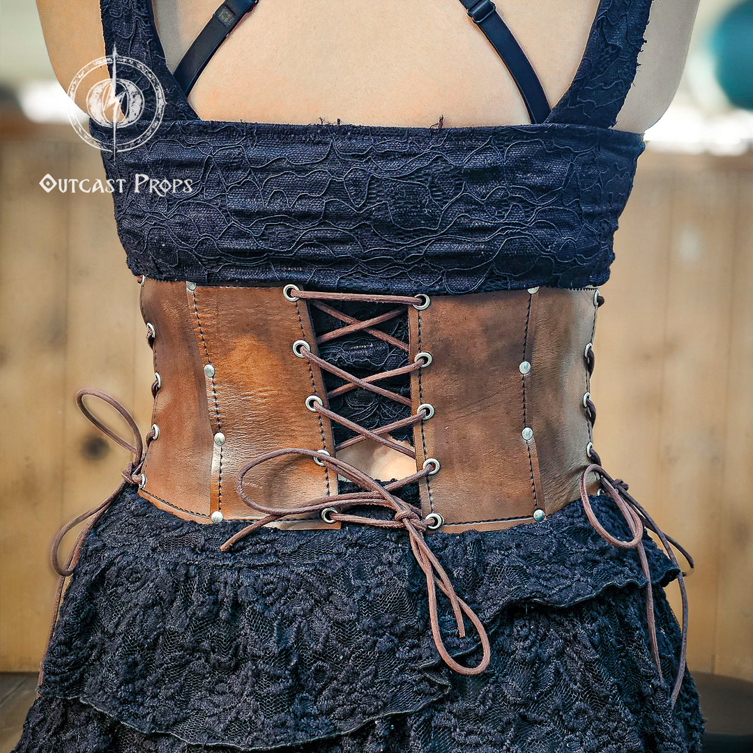 Back view of the aged brown witch leather corset featuring symmetrical lacing for adjustable fit. Made from full-grain vegetable-tanned leather with nickel rivets, this underbust corset enhances gothic and steampunk outfits. Ideal for ren faire costumes or fantasy festivals, it combines historical design and artisan detail. Every Outcast Props corset is hand-aged, making each piece unique in tone and texture.