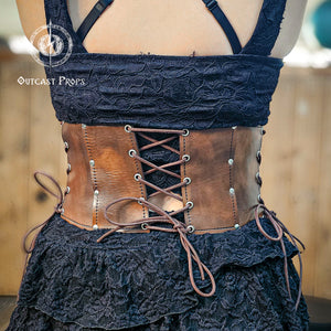 Back view of the aged brown witch leather corset featuring symmetrical lacing for adjustable fit. Made from full-grain vegetable-tanned leather with nickel rivets, this underbust corset enhances gothic and steampunk outfits. Ideal for ren faire costumes or fantasy festivals, it combines historical design and artisan detail. Every Outcast Props corset is hand-aged, making each piece unique in tone and texture.