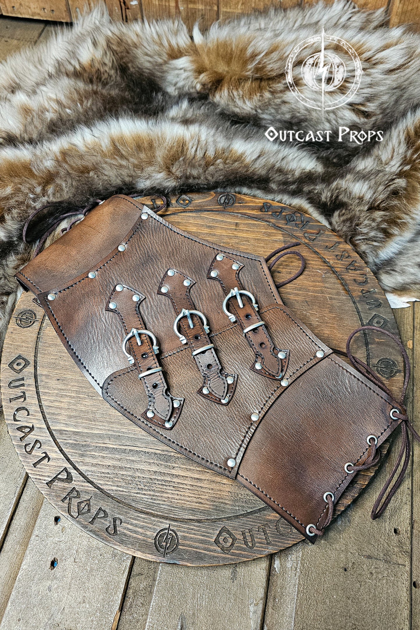 Handcrafted aged brown leather witch corset with nickel hardware displayed on a wooden board surrounded by fur. The waist cincher features triple buckle straps and side lacing, inspired by steampunk and gothic fashion. Ideal for ren faire or LARP costumes, this piece showcases Outcast Props’ signature aging technique and sustainable vegetable-tanned leather.