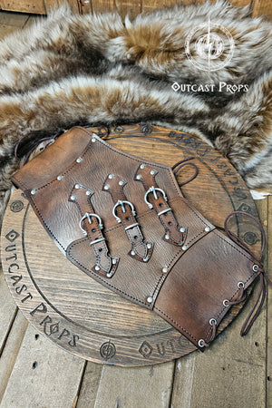 Handcrafted aged brown leather witch corset with nickel hardware displayed on a wooden board surrounded by fur. The waist cincher features triple buckle straps and side lacing, inspired by steampunk and gothic fashion. Ideal for ren faire or LARP costumes, this piece showcases Outcast Props’ signature aging technique and sustainable vegetable-tanned leather.