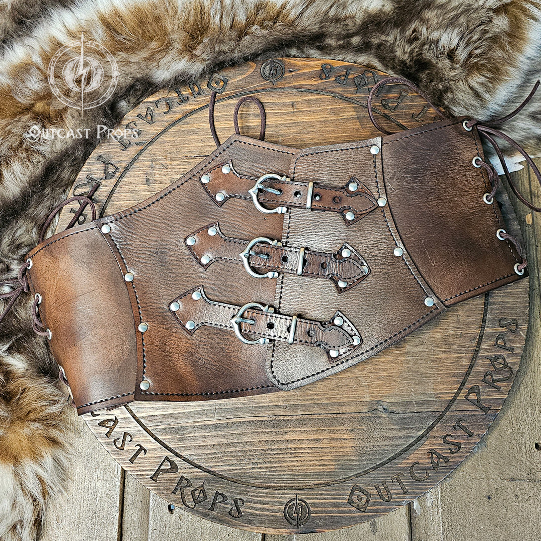 Handcrafted aged brown leather witch corset with nickel hardware displayed on a wooden board surrounded by fur. The waist cincher features triple buckle straps and side lacing, inspired by steampunk and gothic fashion. Ideal for ren faire or LARP costumes, this piece showcases Outcast Props’ signature aging technique and sustainable vegetable-tanned leather.