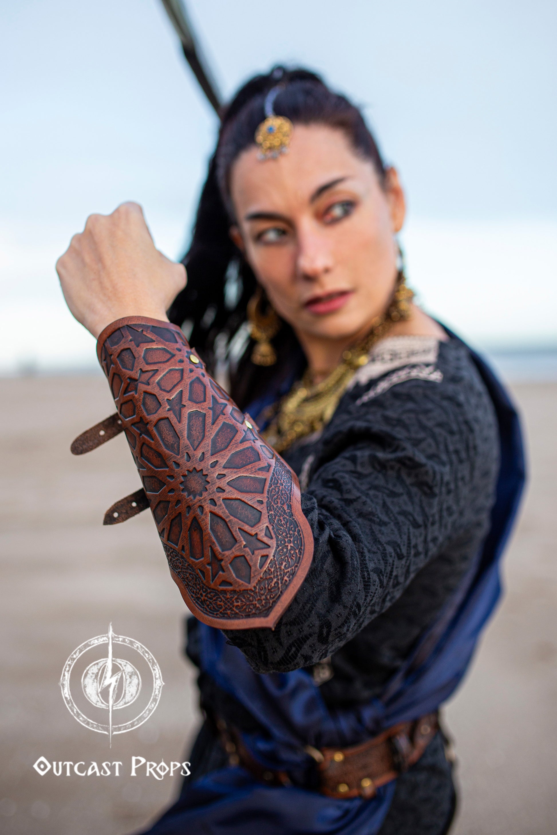 Fantasy female assassin on a beach, wearing brown and blue Arabic pattern leather bracer, part of a medieval stealth outfit with Persian influence. The character holds her arm up, emphasizing the engraved oriental leather bracer suited for LARP, cosplay, and fantasy rogue armor. Her outfit includes rich fabrics and golden jewelry, evoking a mysterious and powerful look. Ideal for players seeking assassin leather bracers with detailed craftsmanship and visual impact.