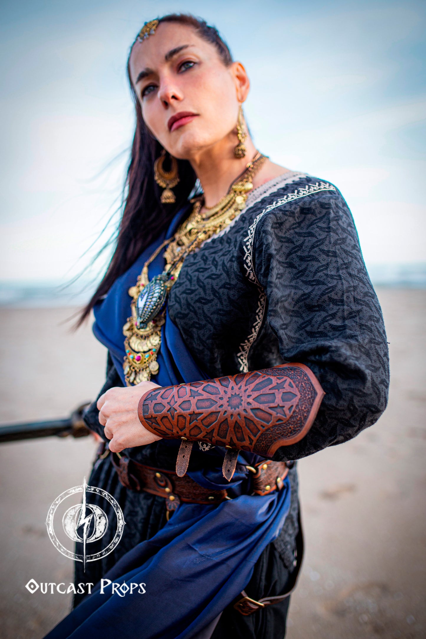 Assassin leather bracers with Arabic pattern in reddish brown worn by a fantasy warrior woman on a beach. She wears ornate jewelry and a dark tunic with a blue sash. The bracers are made from hand-dyed vegetable-tanned leather, engraved with oriental geometric motifs inspired by Alhambra art. Ideal for fantasy rogue armor, stealth LARP costume, or desert-themed cosplay. A perfect accessory for immersive Ren Faire and assassin characters.