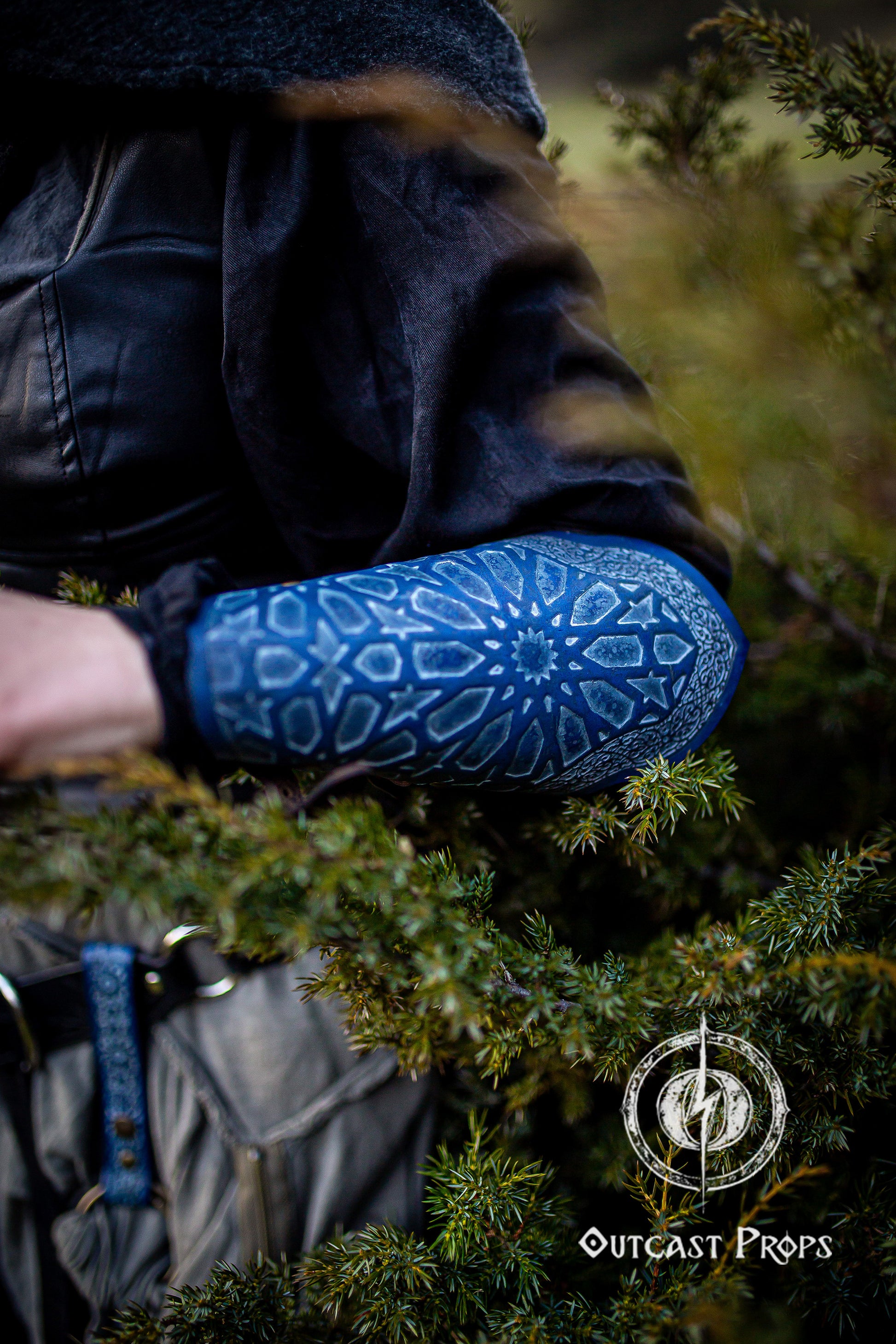 Detailed shot of a blue leather Arabic bracer resting among forest branches, worn over dark layered fabrics and belts. The bracer features engraved geometric motifs inspired by Islamic patterns and dyed with Outcast Props' signature blue. Ideal for fantasy rogue LARP costume, medieval assassin gear, Arabic pattern bracer, stealth cosplay outfit, and oriental fantasy armor for immersive characters moving unseen in woodland or desert scenes.