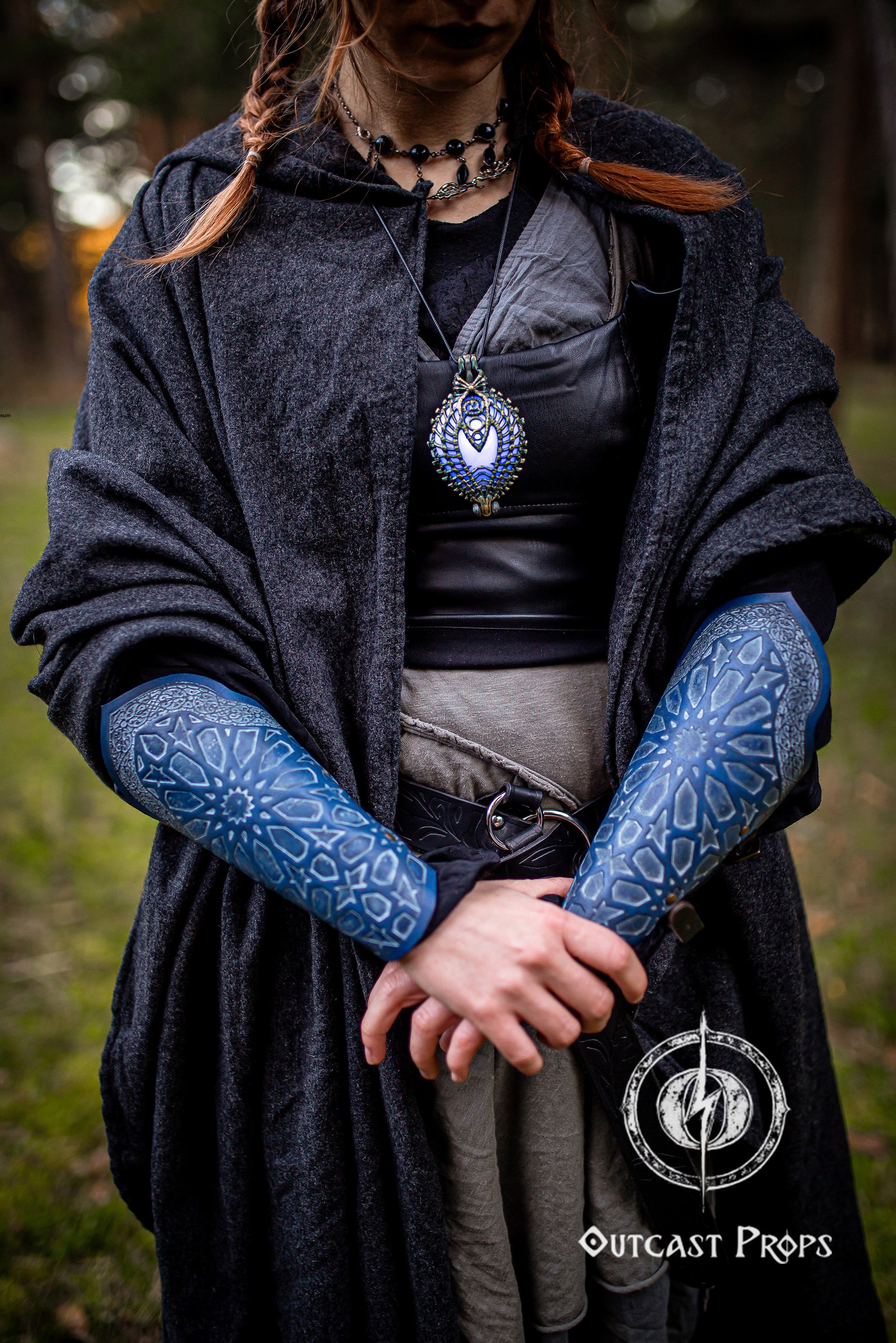 Woman in dark hooded cloak and leather corset standing in the woods, wearing two matching blue engraved leather bracers inspired by Alhambra patterns. Her hands are crossed, showing the full view of the oriental LARP bracers, ideal for stealth characters, assassins, and desert-themed outfits. Her mystical pendant and layered costume enhance the fantasy vibe. These fantasy rogue armor bracers combine comfort, durability, and striking visual impact for cosplay or LARP.