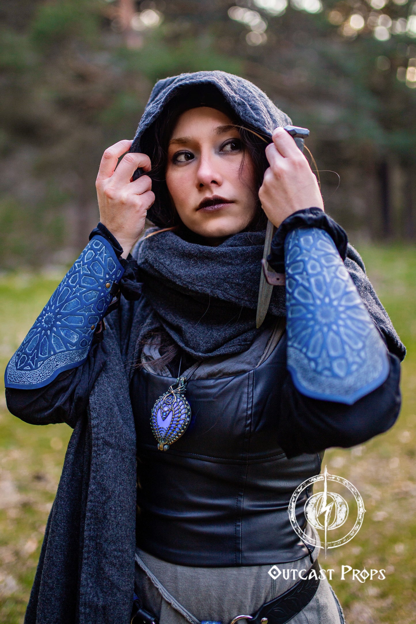 Woman in a hooded gray cloak adjusting her head covering in a forest, wearing matching blue Arabic bracers with engraved geometric patterns. These leather LARP bracers are inspired by Persian architecture and crafted for fantasy rogue or assassin cosplay. Combined with a corset and layered medieval outfit, they complete a stealthy silhouette. Perfect for desert-themed characters, medieval LARP outfits, or cosplay with a nomadic aesthetic.