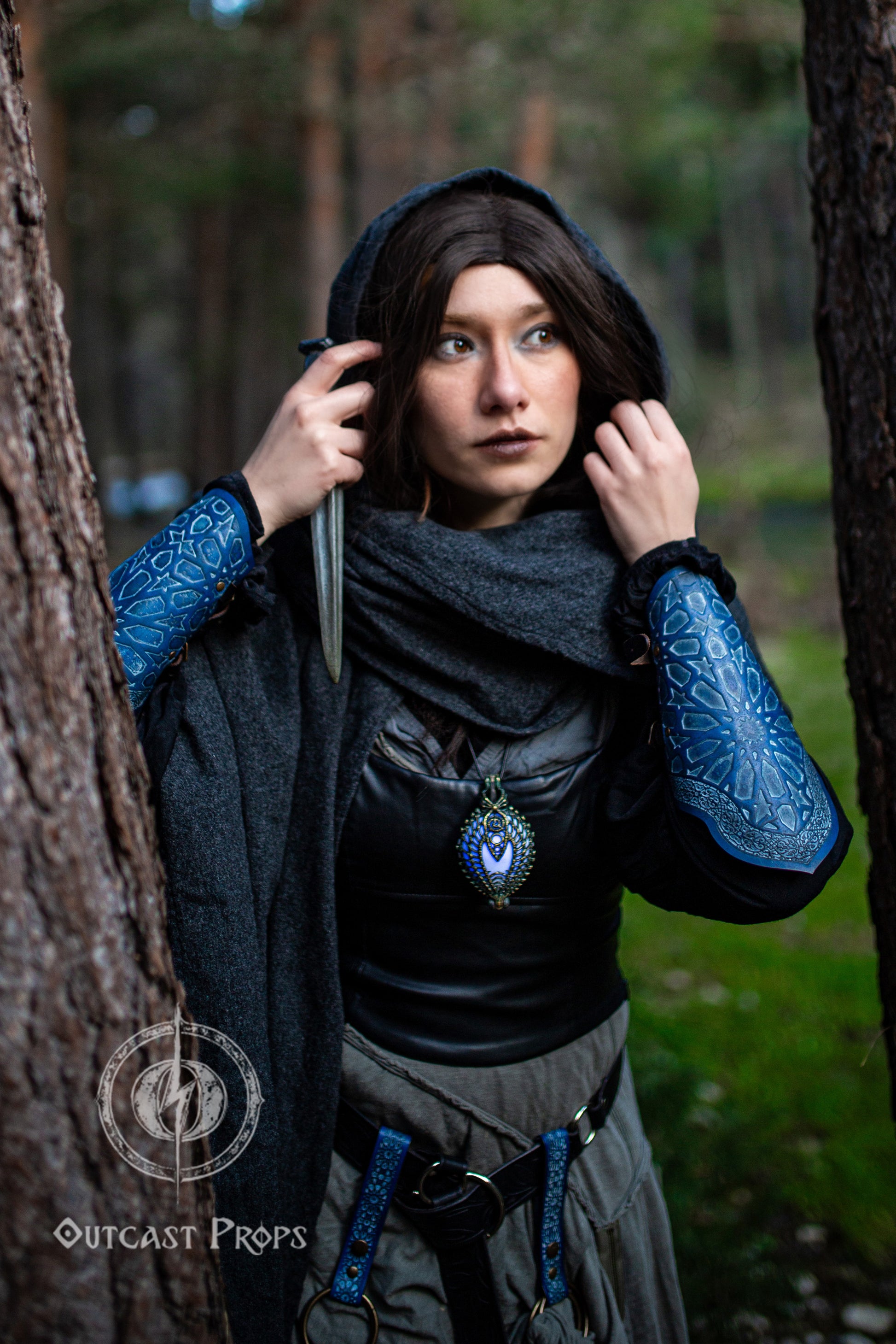 Female LARP rogue standing between trees in a black cloak and gray outfit, showing blue assassin leather bracers with Arabic mosaic pattern. The design is inspired by Alhambra geometry, hand-engraved on vegetable-tanned leather. The bracers pair with a leather corset and matching belt for a stealth costume. Great for fantasy cosplay, oriental rogue gear, or medieval assassin armor. Adjustable and durable for immersive LARP events and desert cosplay kits.
