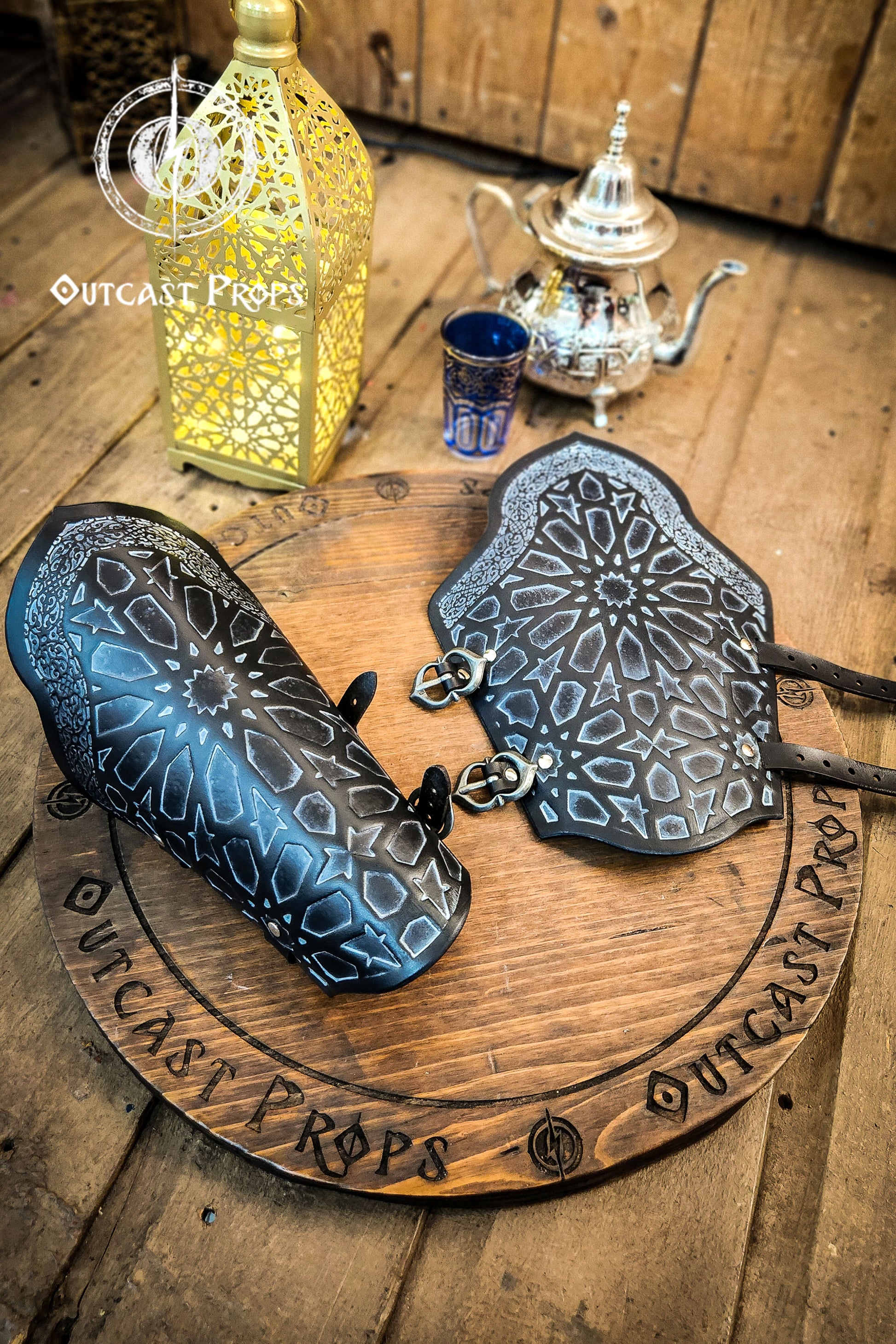 Artistic still life of a pair of black leather bracers with Arabic geometric design, resting on a wooden ritual board surrounded by oriental props like a lantern, tea set, and potion bottles. These assassin leather bracers are ideal for LARP costumes, fantasy rogue armor, and cosplay gear with oriental inspiration. The engraved surface highlights Islamic motifs, while the scene evokes a magical assassin’s den, making the bracers both decorative and practical in use.