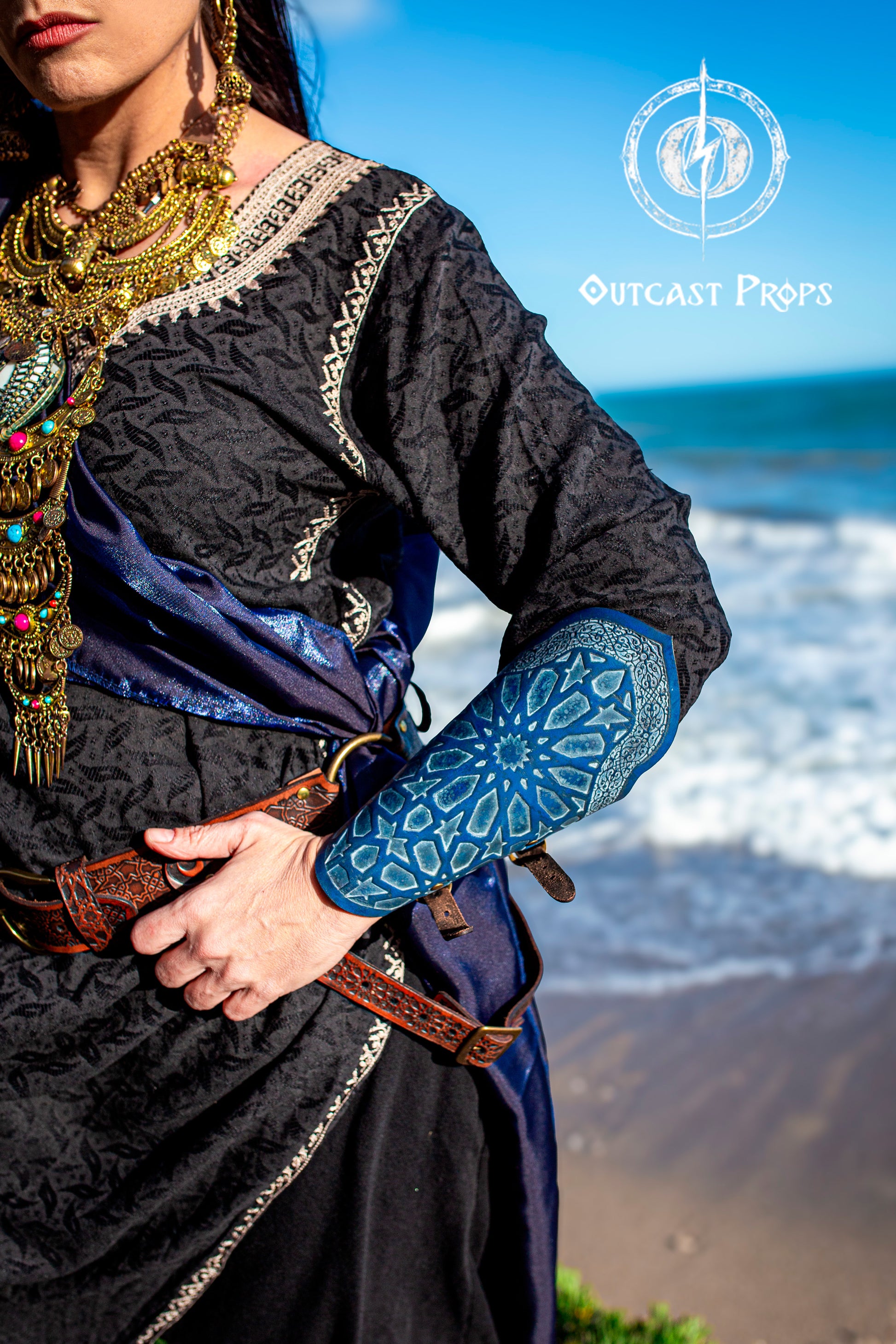Close-up of a blue Assassin Leather Bracer with Arabic pattern worn at the beach, paired with a fantasy rogue outfit featuring ornate jewelry and a double belt. The engraved design recalls Alhambra mosaics, dyed in deep blue and handcrafted for stealth characters. Ideal for LARP bracer blue leather, oriental cosplay arm guard, medieval rogue costume, or assassin fantasy outfit with Arabic armor inspiration and eco leather gear.