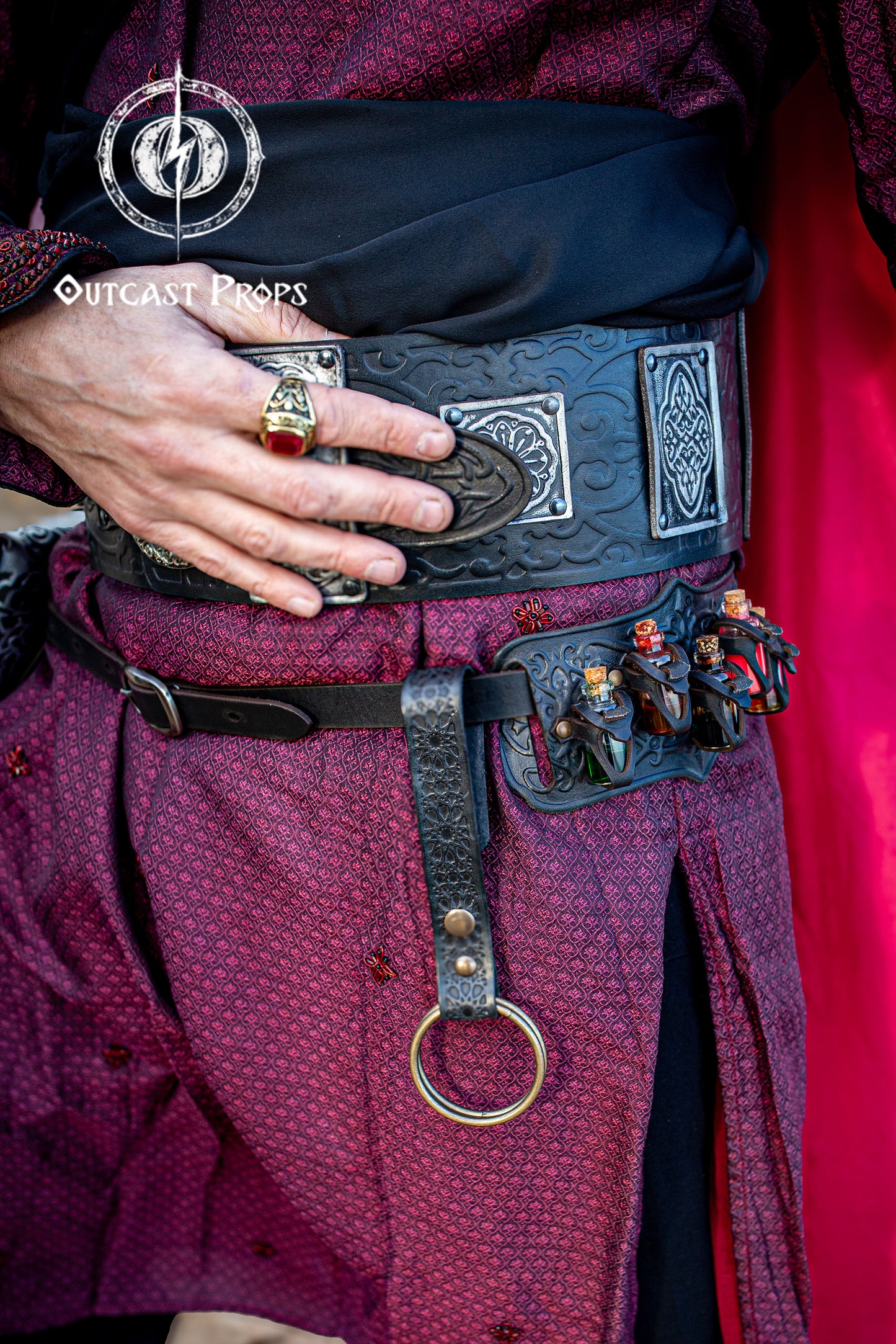 Black medieval Persian leather skirt hike with Alhambra-inspired patterns, worn on an ornate black and silver fantasy belt at a ren faire event. This LARP cosplay accessory features a large brass O-ring and aged snap button, used to hold fabric or suspend items. Perfect for adding practical elegance to renaissance fair costumes, medieval outfits, and fantasy LARP gear, crafted in premium Spanish leather.