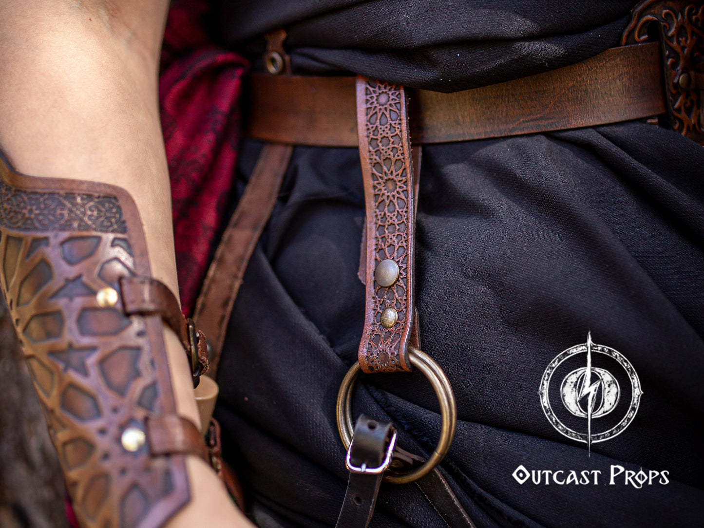 Brown Persian leather skirt hike in close-up, attached to a brown belt and black skirt in a fantasy LARP costume. The handcrafted leather accessory, embossed with Alhambra and Persian medina patterns, features a large brass O-ring for lifting skirts or carrying items. Designed for LARP, renaissance fair, and fantasy cosplay outfits, this skirt hiker adds both visual charm and practical utility to any medieval costume.