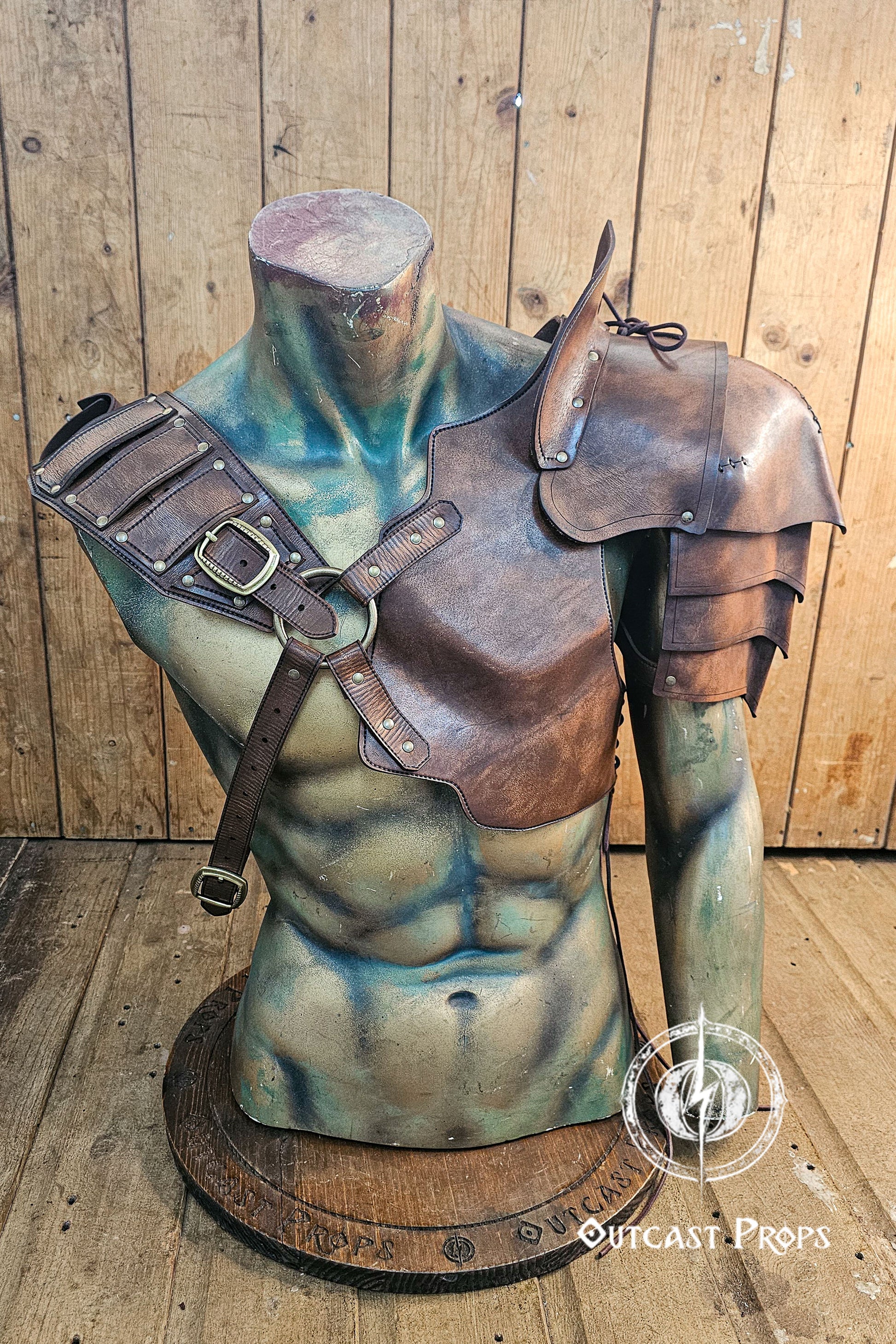 Mannequin bust wearing full barbarian leather shoulder armor set with crossed harness and riveted pauldrons. The armor is aged brown, handcrafted in vegetable-tanned leather with visible patina and hand-stitched details. Worn over a dark medieval shirt, this larp armor set includes sword scabbard on the back. Perfect for viking costume, post-apocalyptic larp, or warrior cosplay outfit.