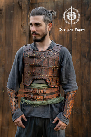 LARP leather armor set in dark brown tones with embossed battle scars and antique brass rivets. The warrior wears a full leather torso armor with triple buckle closure and matching leather vambraces. Designed for barbarian cosplay, fantasy roleplay and renfaire costumes, this handcrafted leather armor features Outcast Props' signature aging technique and embossed panels evoking a raw, tribal look. Viking Studded Leather Armor Chest Armor for Men LARP Armor Witcher Leather Armour Medieval Leather Armor