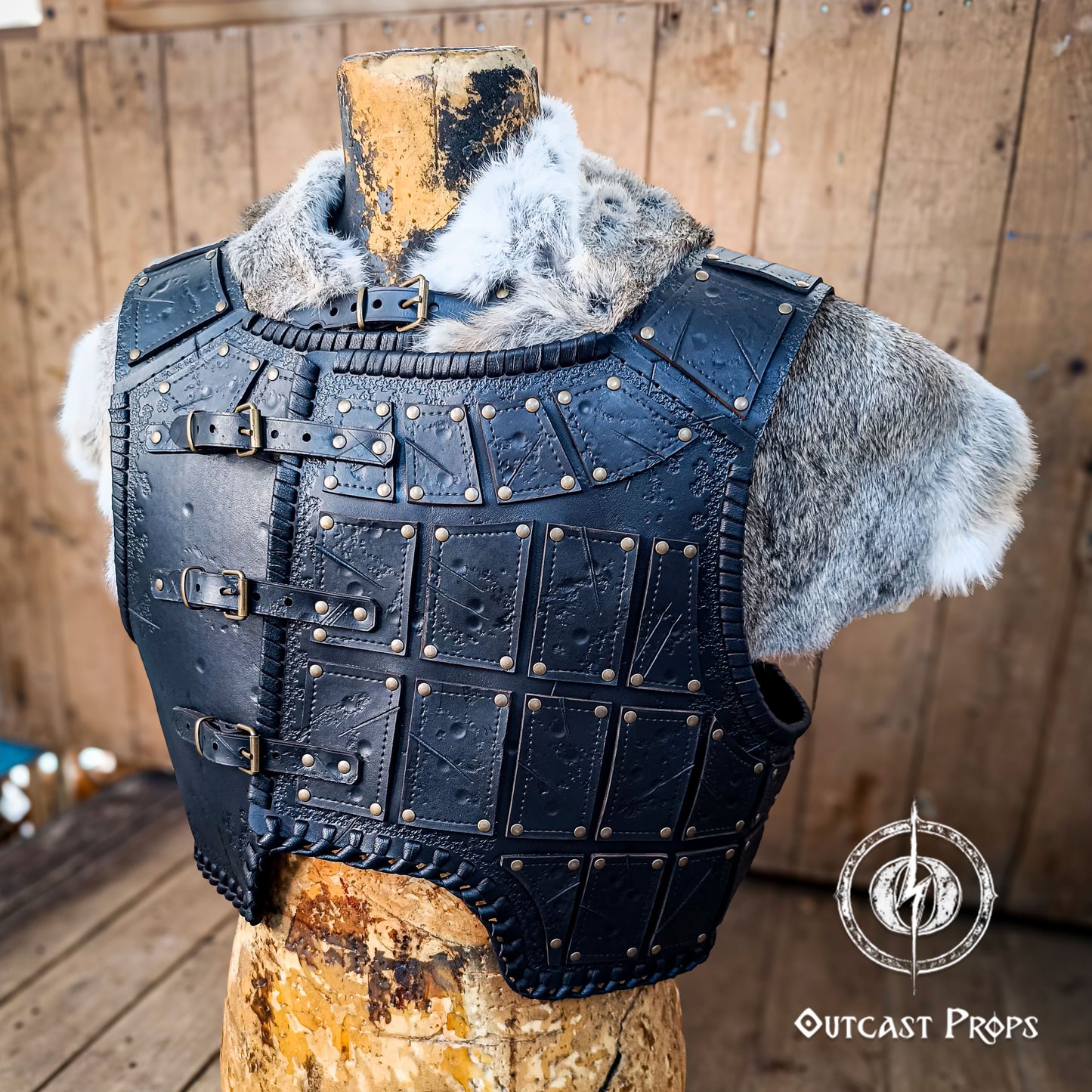 A handmade leather studded armor displayed on a wooden stand with a fur cape, featuring brown leather plates with studs, and a corded edge finish. Barbarian studded leather chest armor with rabbit fur cape attachment shown on mannequin. Perfect for fantasy LARP, Viking reenactment or cosplay, this full-grain leather armor is aged by hand and paired here with fur for an epic look. Adjustable buckle straps and visible stitching add to the rugged charm of this high-impact warrior costume piece.