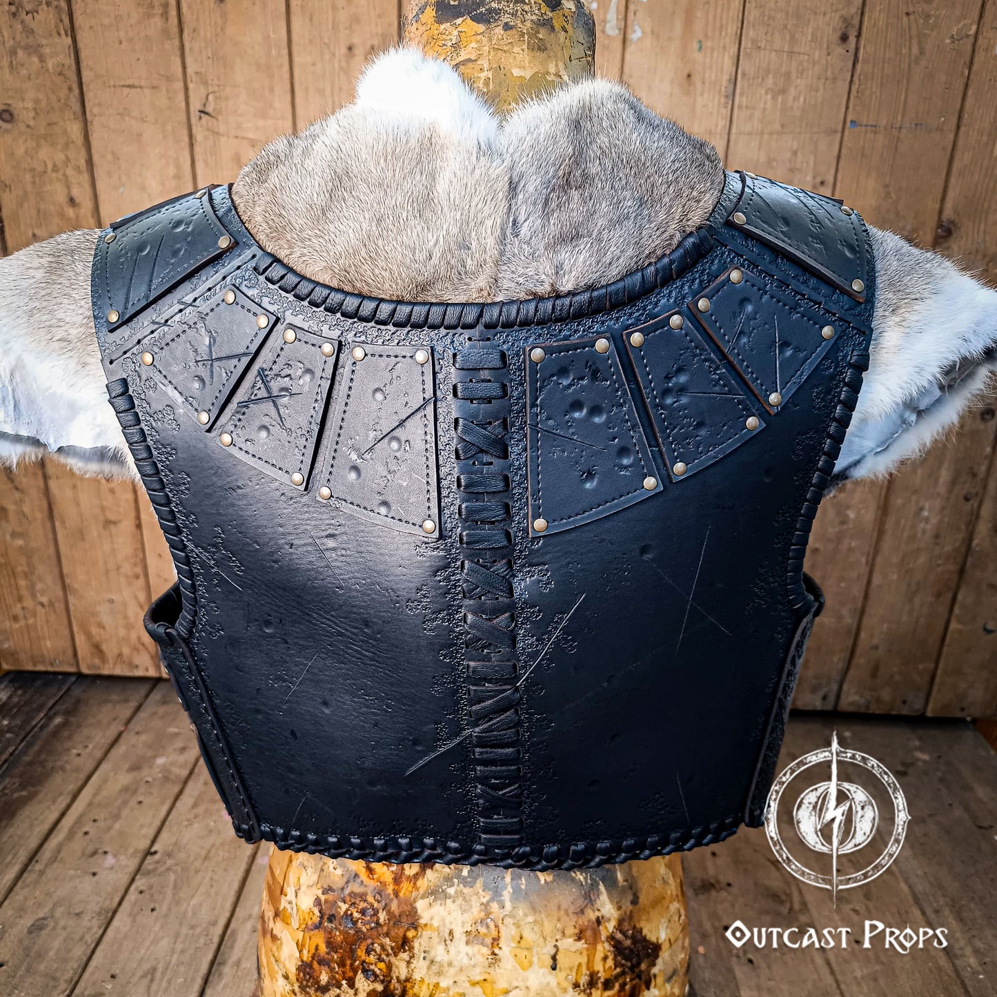 Black leather armor vest with fur lining on a wooden stand, branded 'Outcast Props'. A handmade leather studded armor displayed on a wooden stand with a fur cape, featuring brown leather plates with studs, and a corded edge finish. Barbarian studded leather chest armor with rabbit fur cape attachment shown on mannequin. Perfect for fantasy LARP, Viking reenactment or cosplay, this full-grain leather armor is aged by hand and paired here with fur for an epic look. Adjustable buckle straps and visible stitchi