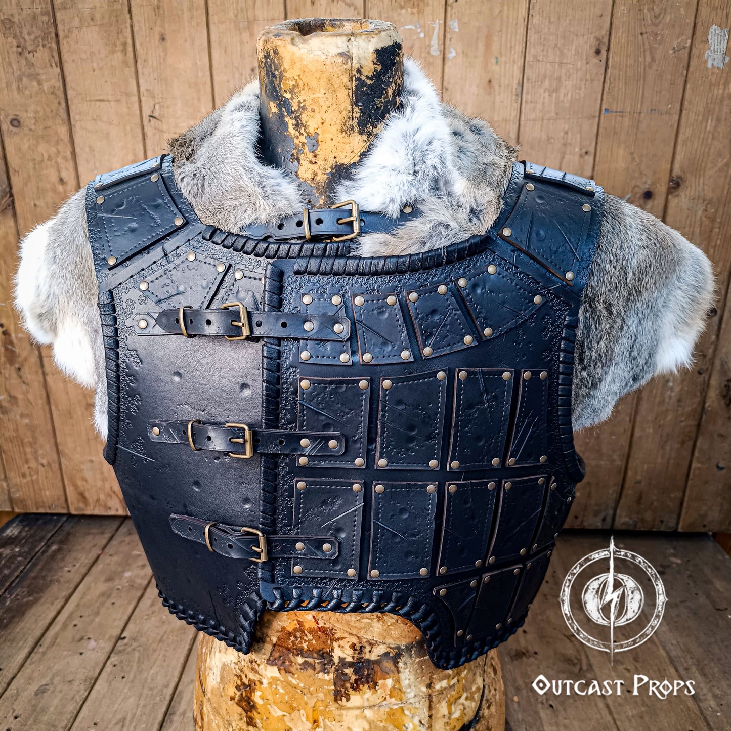 Viking-style leather armor on a wooden stand with Outcast Props logo. A handmade leather studded armor displayed on a wooden stand with a fur cape, featuring brown leather plates with studs, and a corded edge finish. Barbarian studded leather chest armor with rabbit fur cape attachment shown on mannequin. Perfect for fantasy LARP, Viking reenactment or cosplay, this full-grain leather armor is aged by hand and paired here with fur for an epic look. Adjustable buckle straps and visible stitching