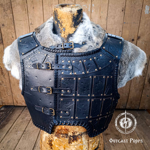 Viking-style leather armor on a wooden stand with Outcast Props logo. A handmade leather studded armor displayed on a wooden stand with a fur cape, featuring brown leather plates with studs, and a corded edge finish. Barbarian studded leather chest armor with rabbit fur cape attachment shown on mannequin. Perfect for fantasy LARP, Viking reenactment or cosplay, this full-grain leather armor is aged by hand and paired here with fur for an epic look. Adjustable buckle straps and visible stitching