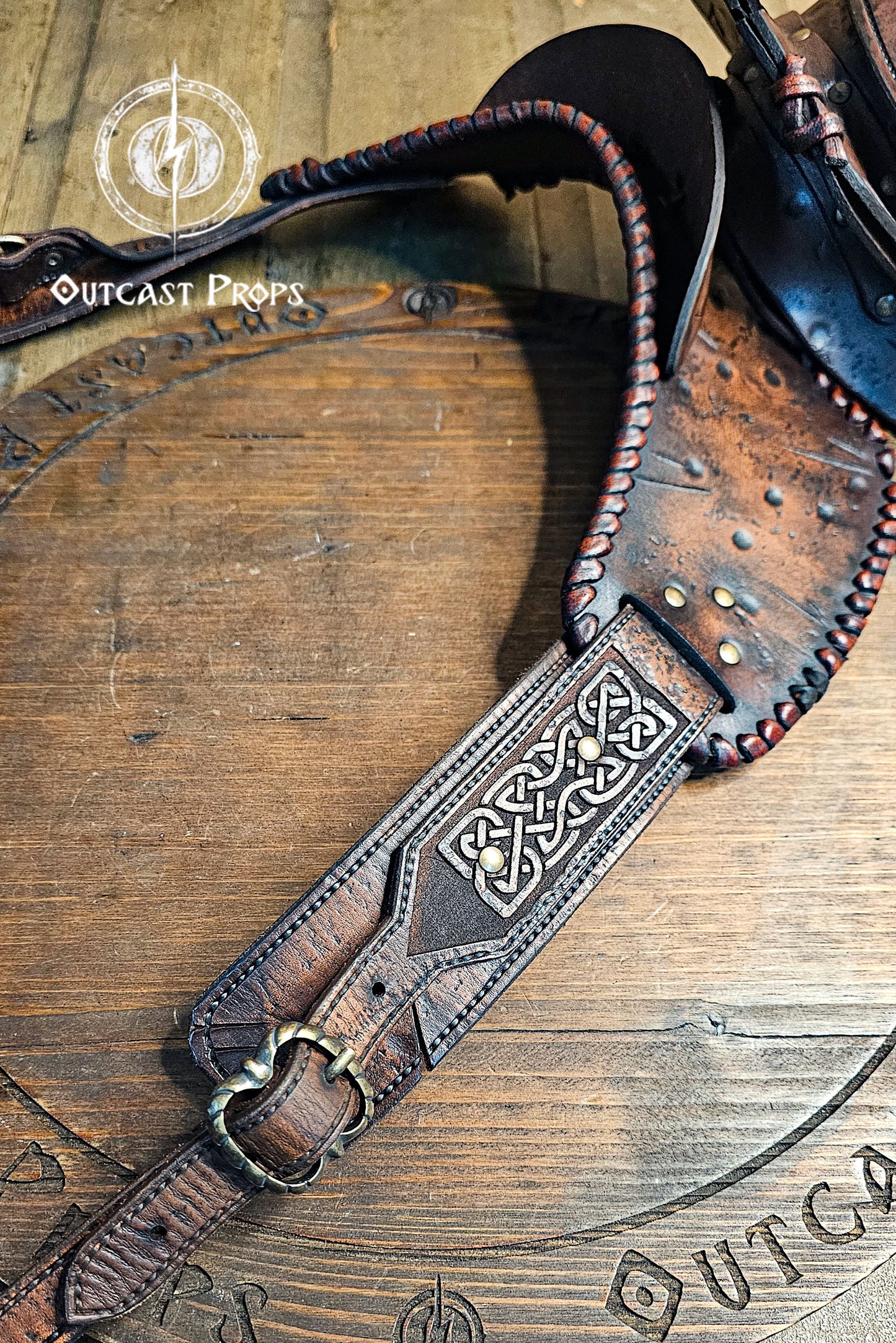 Barbarian Viking Leather Shoulder armor set. Brown leather strap with intricate design on a wooden surface, featuring 'Outcast Props' branding.