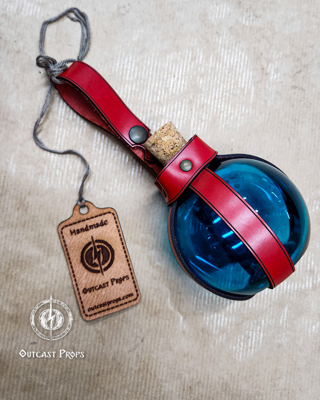 Blue spherical object with red leather strap and cork stopper on a beige background, featuring 'Outcast Props' branding. Potion Bottle with Leather Holder. Safe Flask DRINKING Bottle for Larp, Costume and Cosplay. Alchemist, Steampunk. No crystal. Made of PET