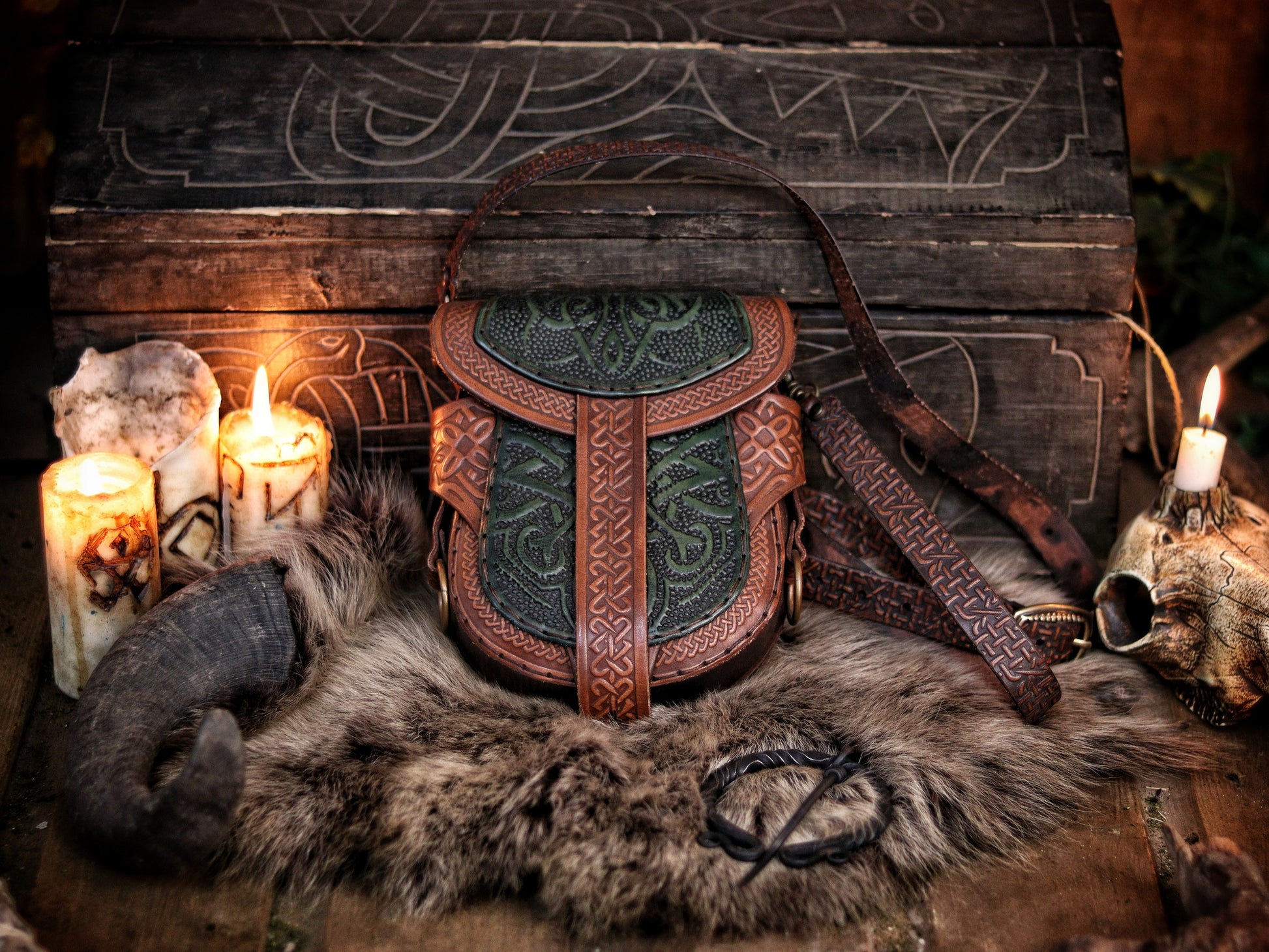 Celtic leather bag with embossed knotwork and green panels, surrounded by candles, a carved wooden chest, and fur. This medieval belt pouch features rich texture and earthy tones, ideal for LARP, ren faire, or fantasy cosplay. The embossed design and aged finish highlight the artisanal craftsmanship. Strap and belt loops offer versatile wear. Premium leather accessory for Viking and Celtic characters.