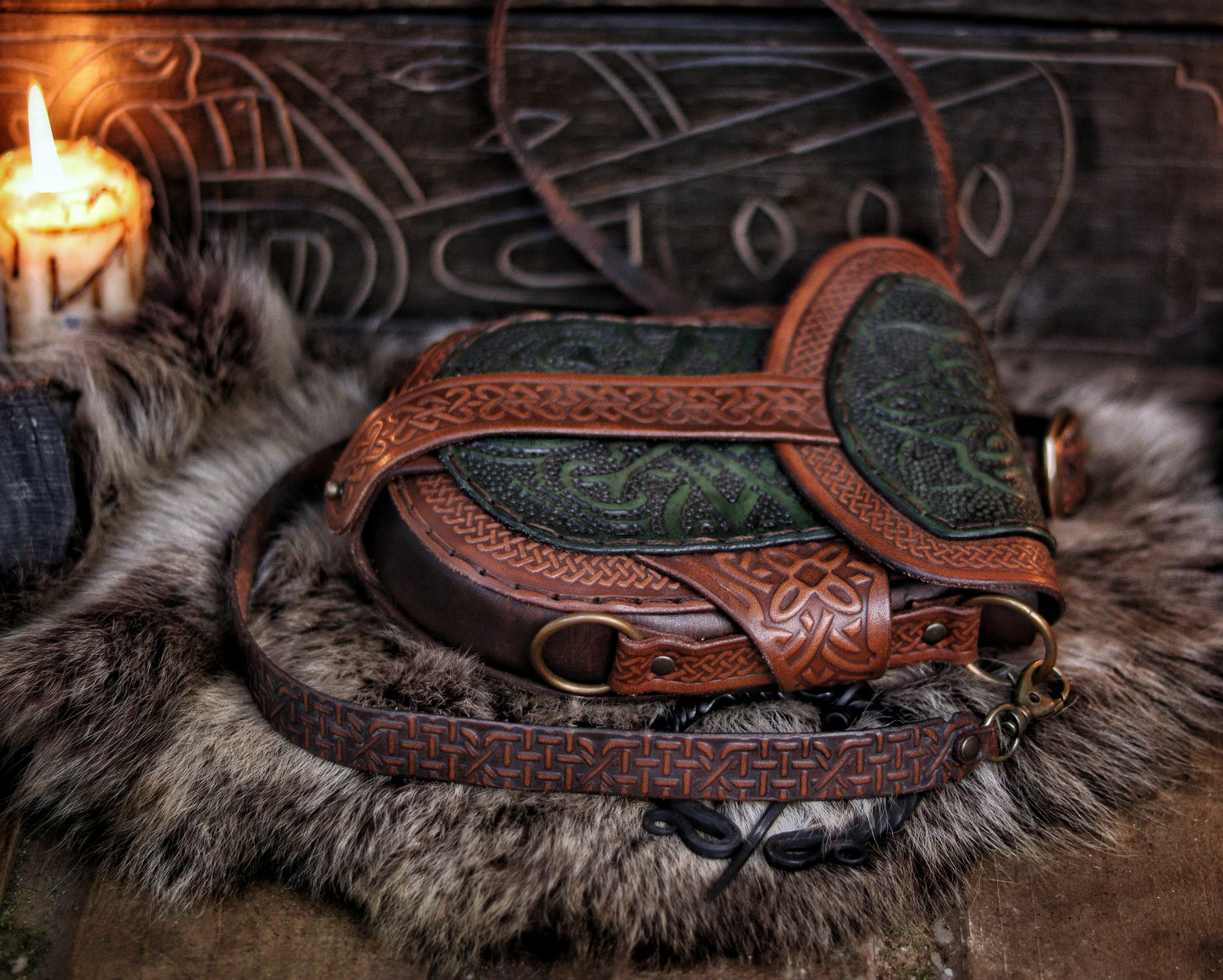 Close-up side view of a green and brown Celtic leather bag resting on fur, showing embossed details and engraved strap. The handcrafted bag blends traditional knotwork with a rustic fantasy look, perfect for Renaissance fair or LARP use. Vegetable-tanned leather and layered textures offer elegance and durability. Great for costume use or daily wear with medieval flair. Strap rings and loops visible
