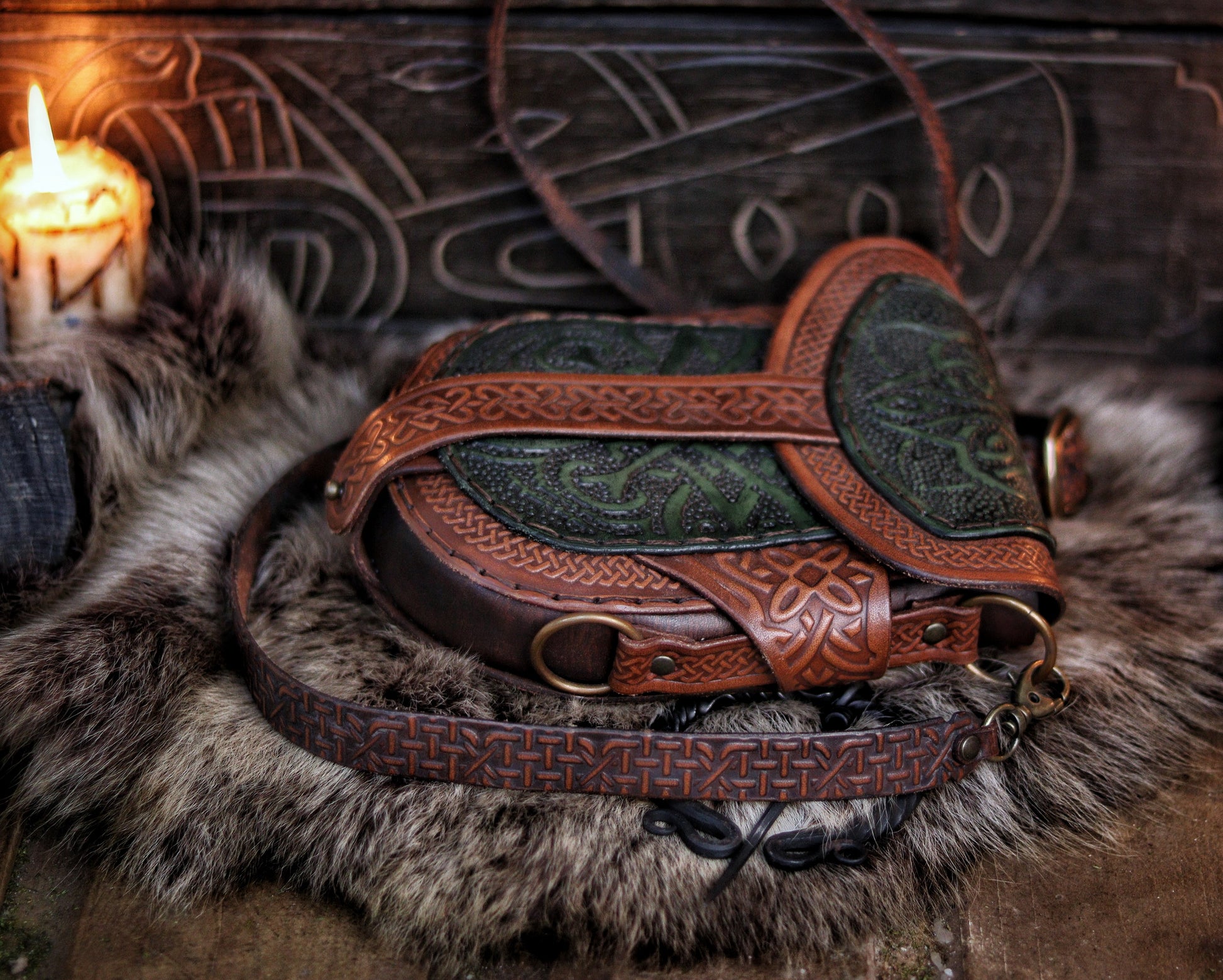 Close-up side view of a green and brown Celtic leather bag resting on fur, showing embossed details and engraved strap. The handcrafted bag blends traditional knotwork with a rustic fantasy look, perfect for Renaissance fair or LARP use. Vegetable-tanned leather and layered textures offer elegance and durability. Great for costume use or daily wear with medieval flair. Strap rings and loops visible