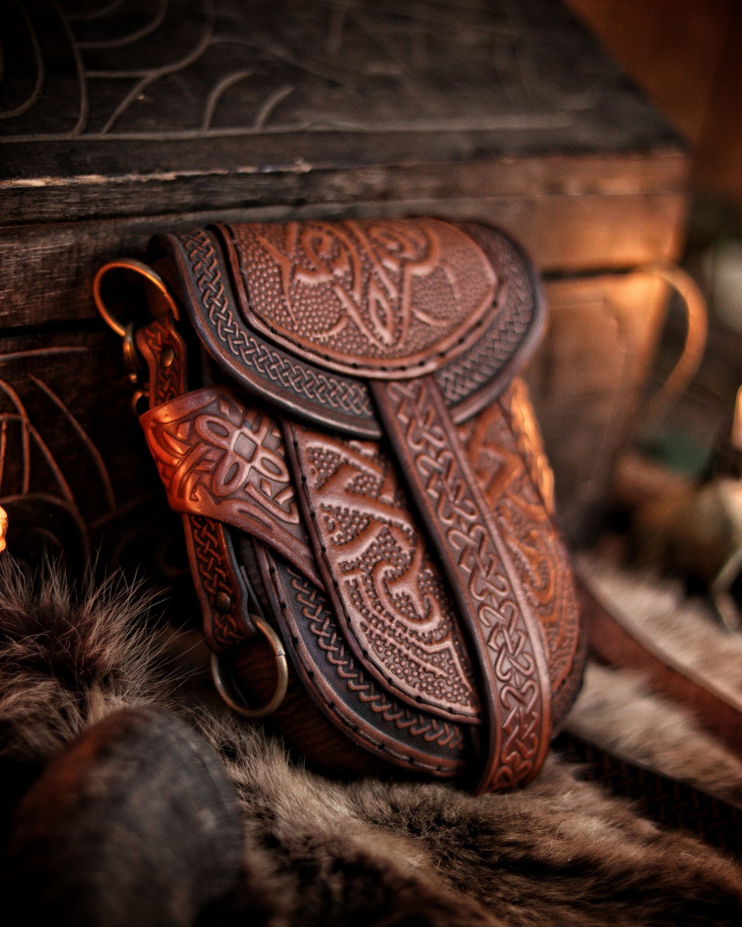 Brown Celtic pouch with hand-embossed leather, placed against rustic wood and soft fur. The intricate knotwork and flame-like embossing evoke fantasy and ancient warrior aesthetics. Ideal for LARP costumes, renaissance fair outfits, or as a daily medieval accessory. Handcrafted in Spain using sustainable materials, with dual carrying options. Perfect blend of beauty and function.