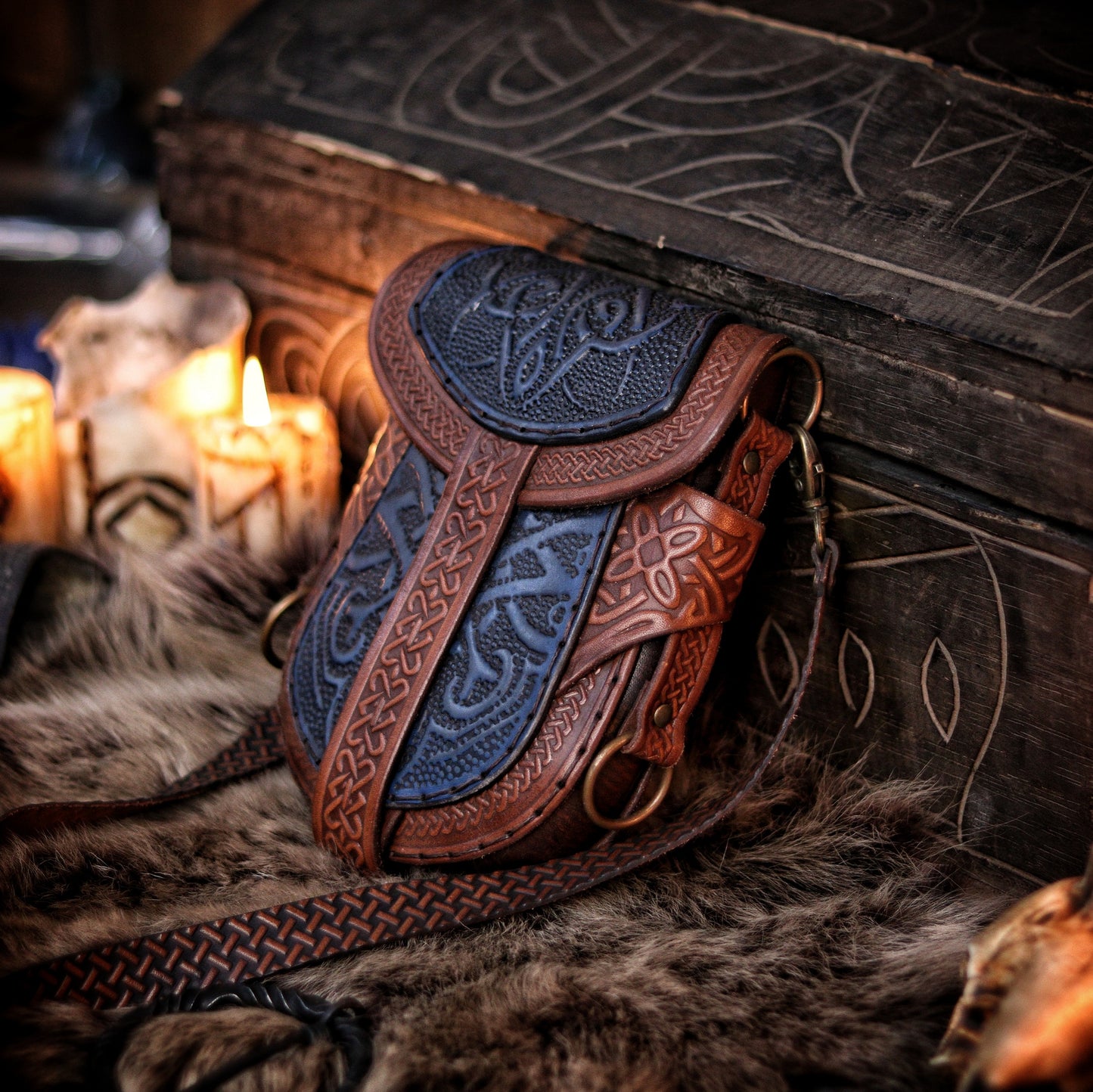 Blue and brown Celtic leather bag displayed upright beside candles and fur, in front of a carved wooden chest. Embossed with knotwork and aged by hand, this fantasy belt pouch is ideal for Viking, Celtic, or medieval LARP outfits. The structure allows both shoulder strap and belt attachment. Perfect size for essentials like phone and wallet. Premium full-grain leather with painted two-tone finish.