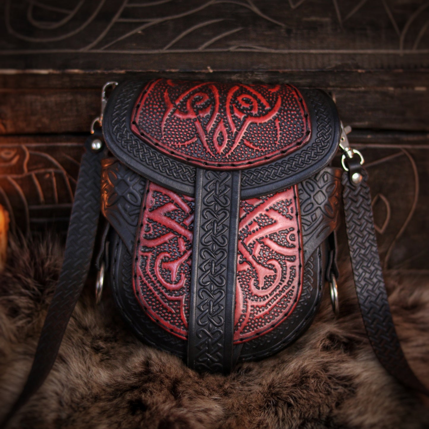 Close-up frontal view of red and black Celtic leather bag with symmetrical embossed designs. The pouch rests on fur with a textured leather strap on both sides. This piece offers premium craftsmanship and bold design for Viking or fantasy characters. Practical and eye-catching, it’s ideal for carrying small items at events or everyday medieval-themed style. Durable, adjustable, and wearable.