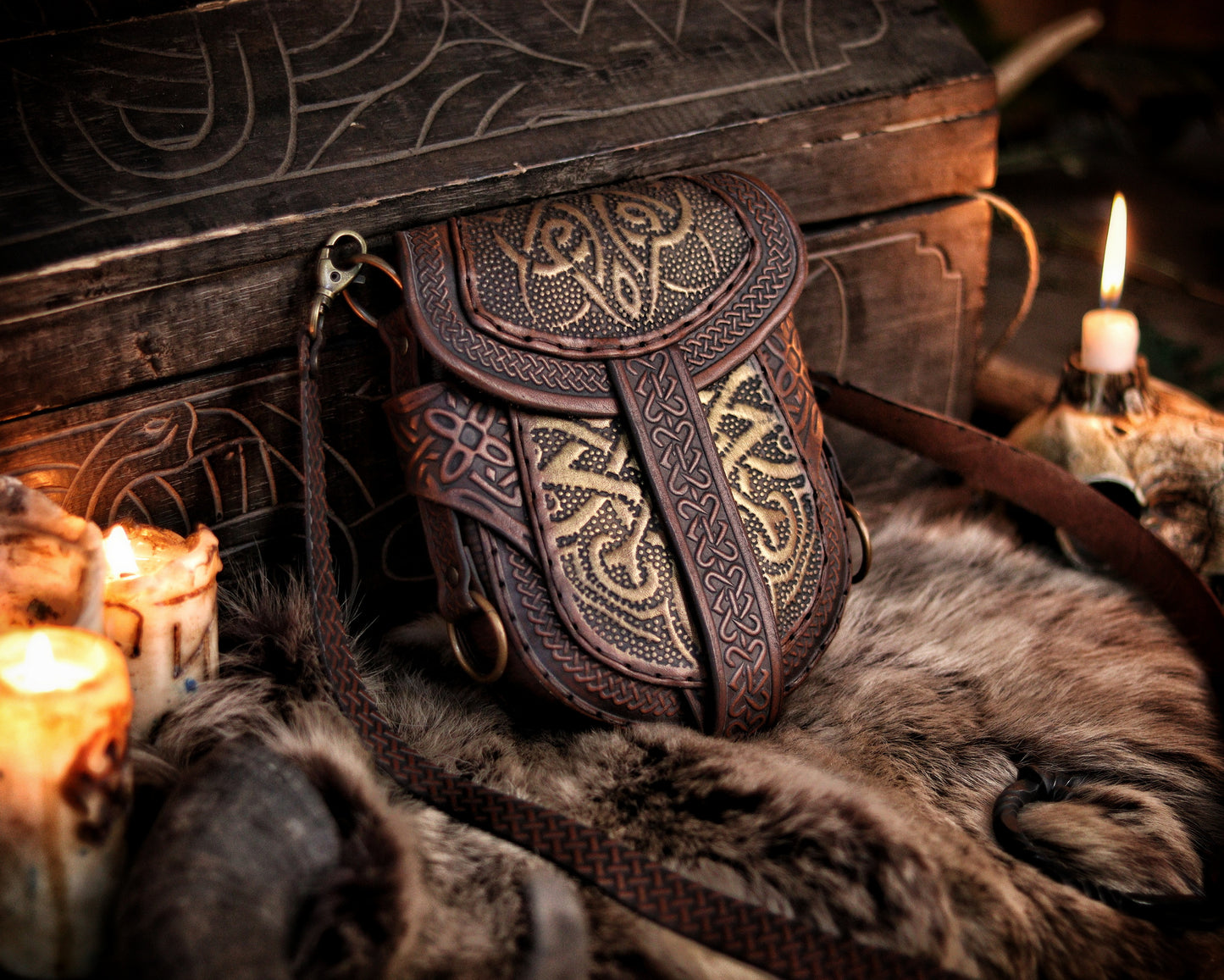 Brown and gold Celtic leather bag displayed upright beside candles and fur, in front of a carved wooden chest. Embossed with knotwork and aged by hand, this fantasy belt pouch is ideal for Viking, Celtic, or medieval LARP outfits. The structure allows both shoulder strap and belt attachment. Perfect size for essentials like phone and wallet. Premium full-grain leather with painted two-tone finish.