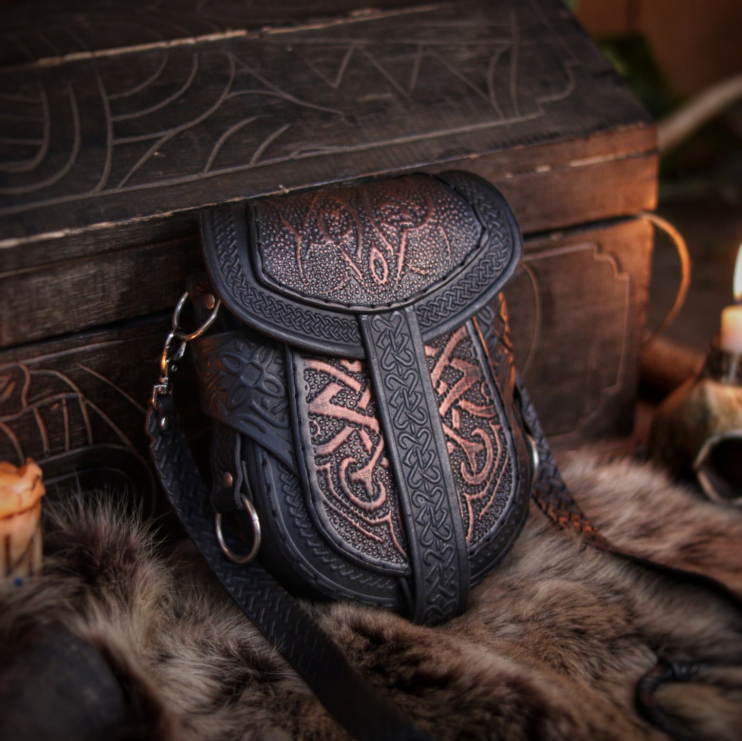 Black and bronze Celtic leather bag displayed upright beside candles and fur, in front of a carved wooden chest. Embossed with knotwork and aged by hand, this fantasy belt pouch is ideal for Viking, Celtic, or medieval LARP outfits. The structure allows both shoulder strap and belt attachment. Perfect size for essentials like phone and wallet. Premium full-grain leather with painted two-tone finish.