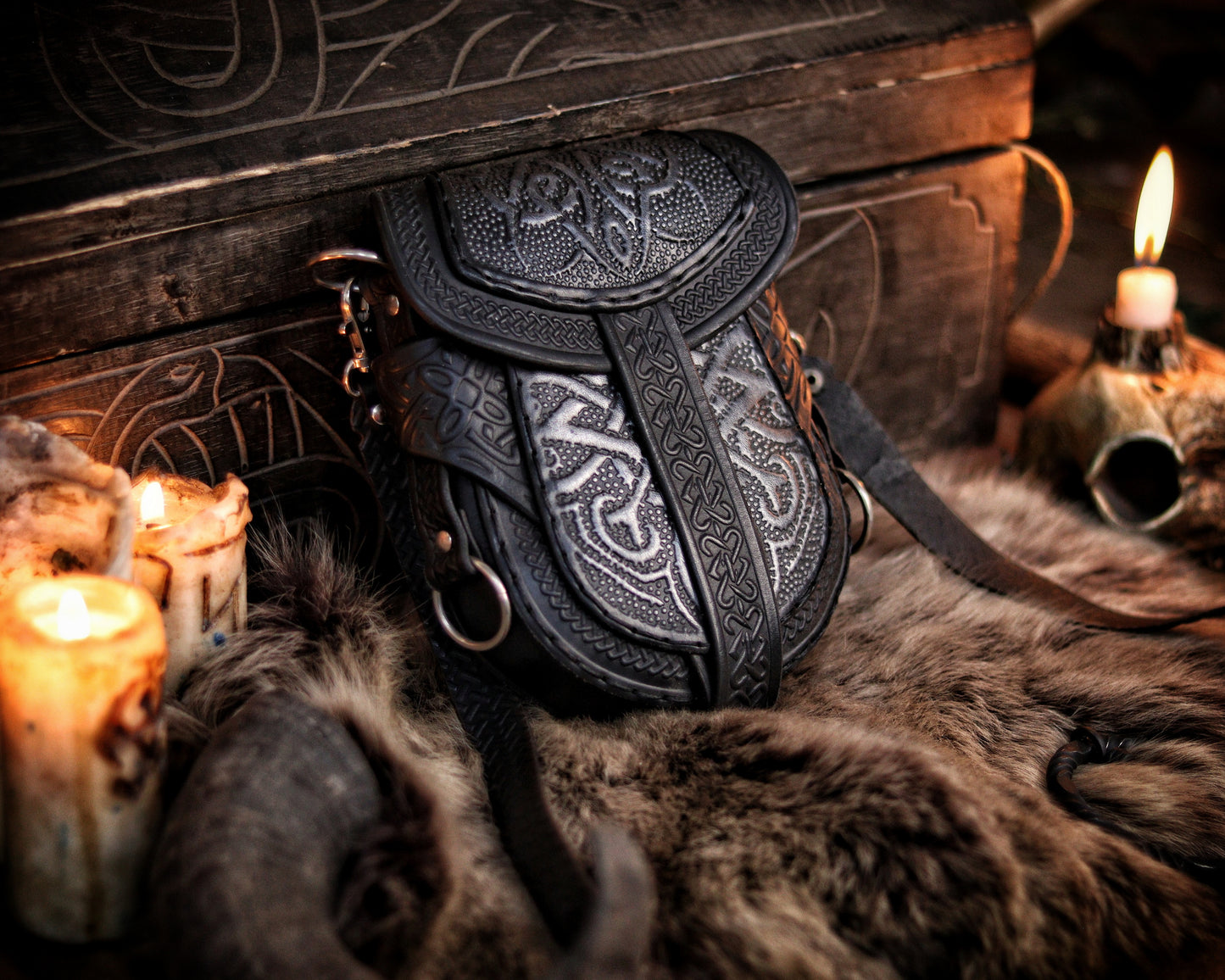 Black and silver Celtic leather bag displayed upright beside candles and fur, in front of a carved wooden chest. Embossed with knotwork and aged by hand, this fantasy belt pouch is ideal for Viking, Celtic, or medieval LARP outfits. The structure allows both shoulder strap and belt attachment. Perfect size for essentials like phone and wallet. Premium full-grain leather with painted two-tone finish.