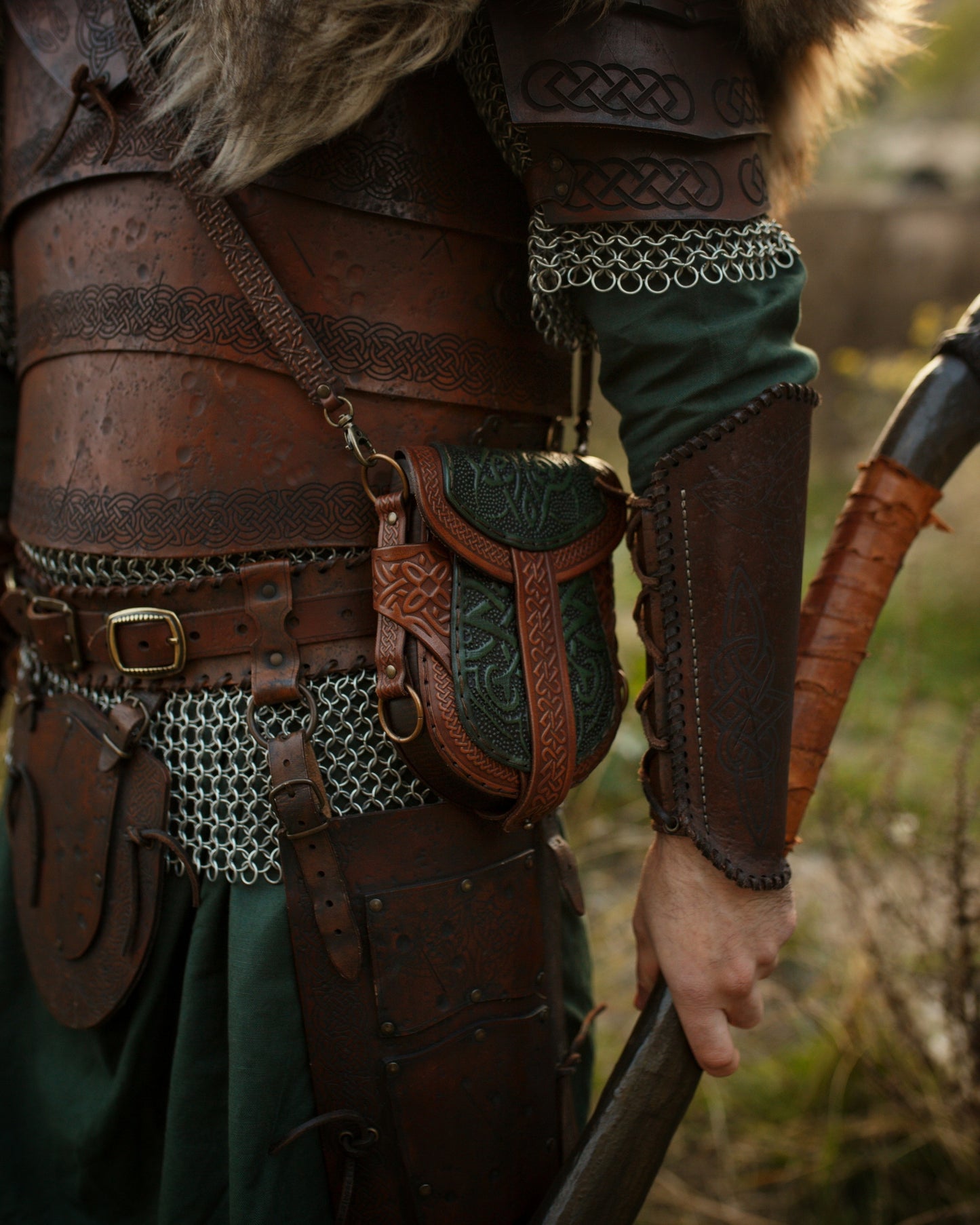 Green and brown Celtic leather bag worn on the hip of a warrior in full armor. The bag hangs from a belt and matches the handcrafted leather armor. Designed for LARP or reenactment, it holds essentials securely and complements fantasy or historical costumes. The embossed design stands out with natural dyes and a battle-worn finish. Ideal for roleplayers who value utility and style.