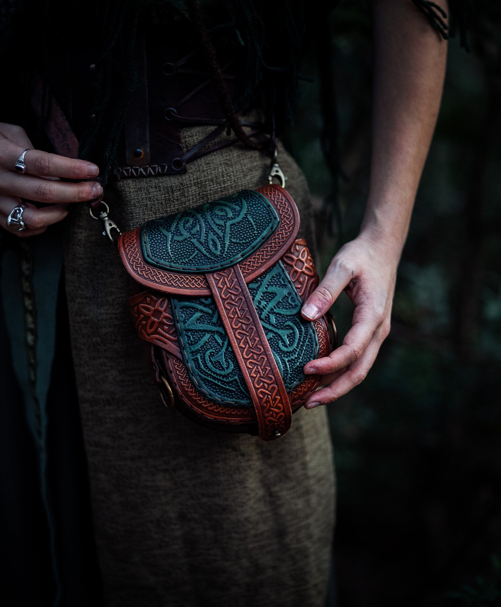 Close-up of a Celtic leather pouch held by both hands. The green embossed panels and brown leather frame create a rich fantasy look. The detailed craftsmanship makes it ideal for immersive LARP characters or medieval-themed daily wear. The pouch is secured with a leather strap and can be worn with or without a shoulder strap. A standout accessory for anyone seeking authenticity and elegance.